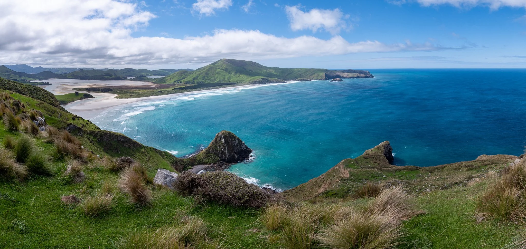 A scenic view of a coastline with green hills, a sandy beach, and blue ocean waves, under a partly cloudy sky.