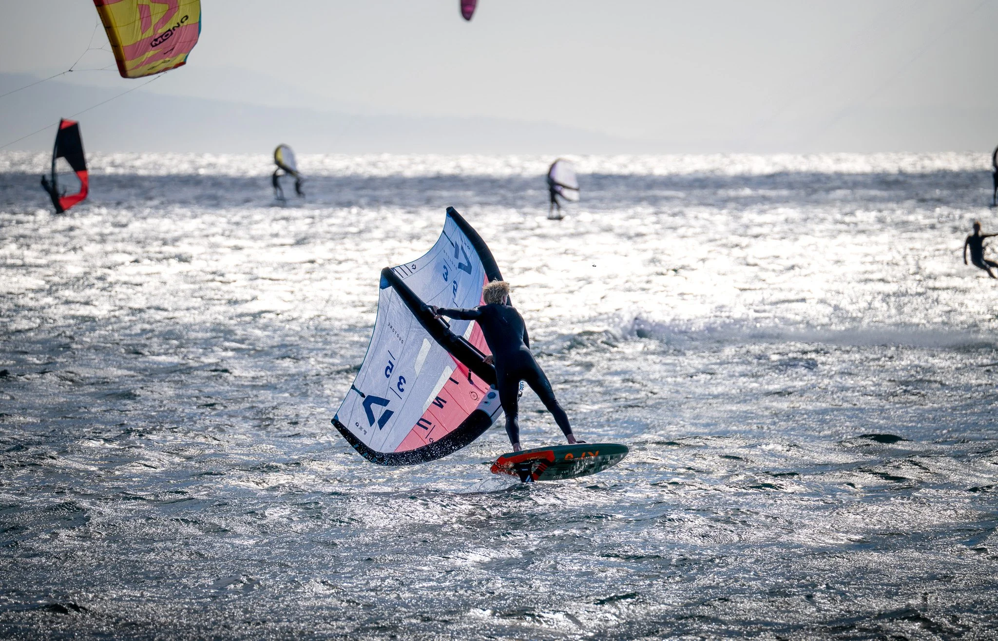 Person Wingfoiling, performing a technical manoeuvre on brightly lit water with other windsurfers in the distance.