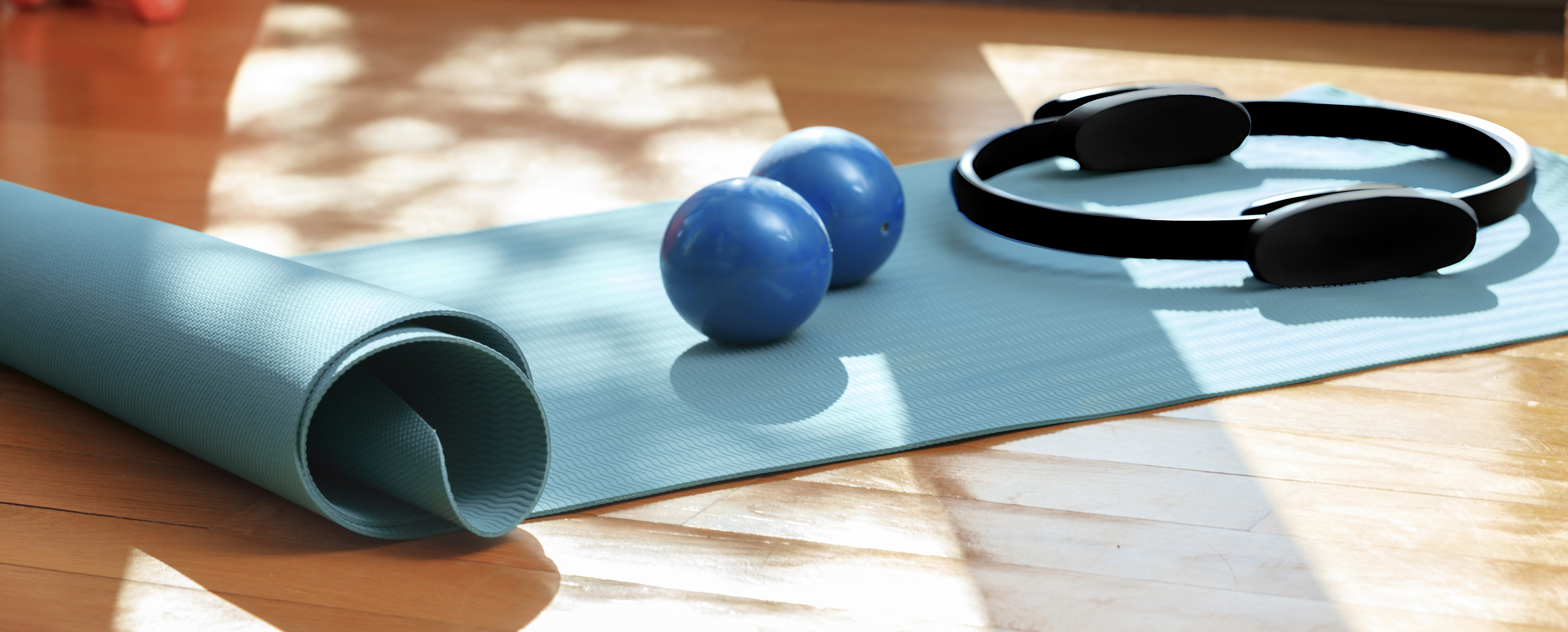 A light blue pilates mat is rolled on a wooden floor, with two dark blue exercise balls and a black pilates ring resting on it. Sunlight casts shadows across the mat.