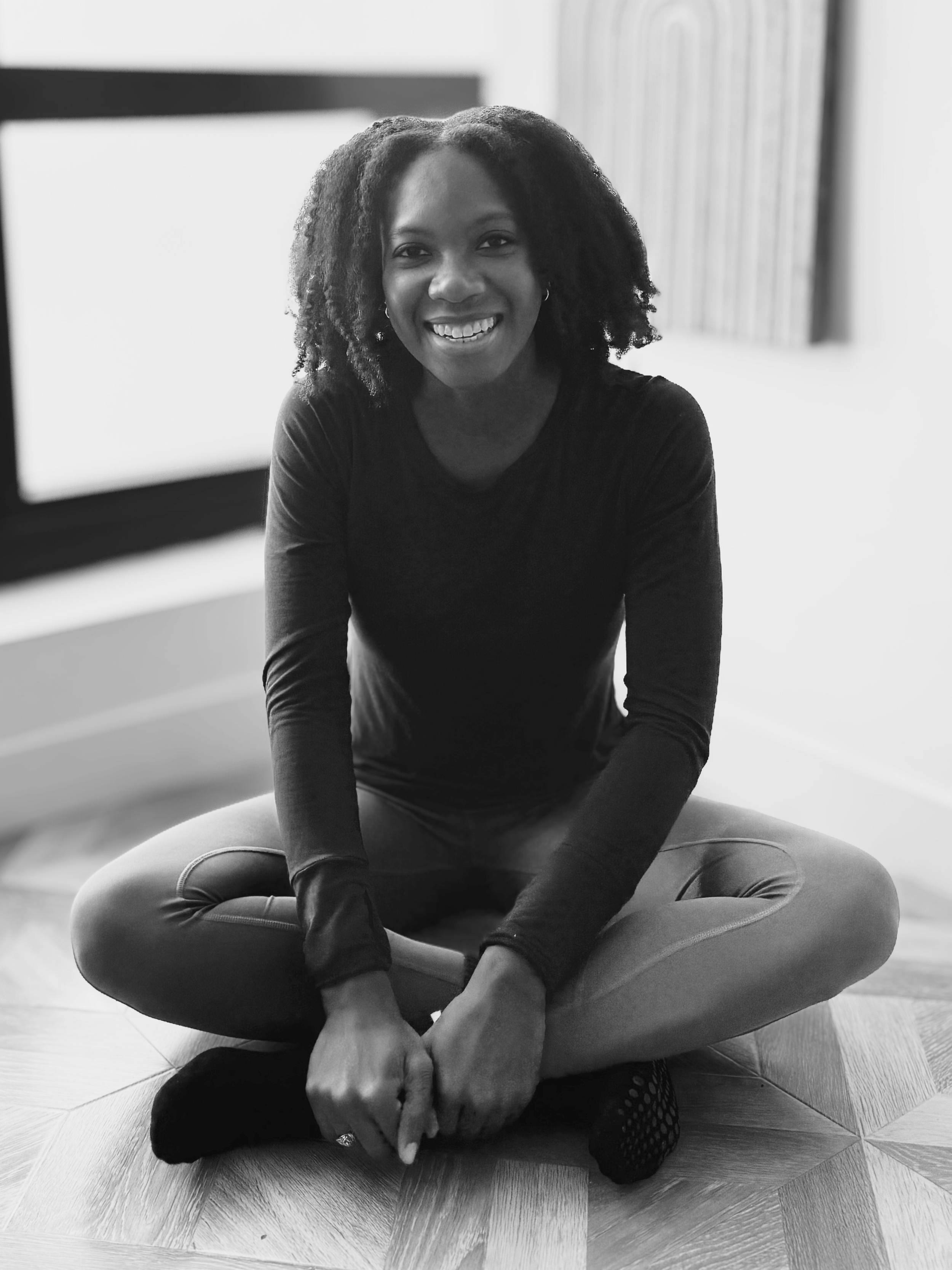 Courtney, founder of True Align Pilates, wearing a long-sleeved shirt and leggings siting cross-legged on a wooden floor and smiling.