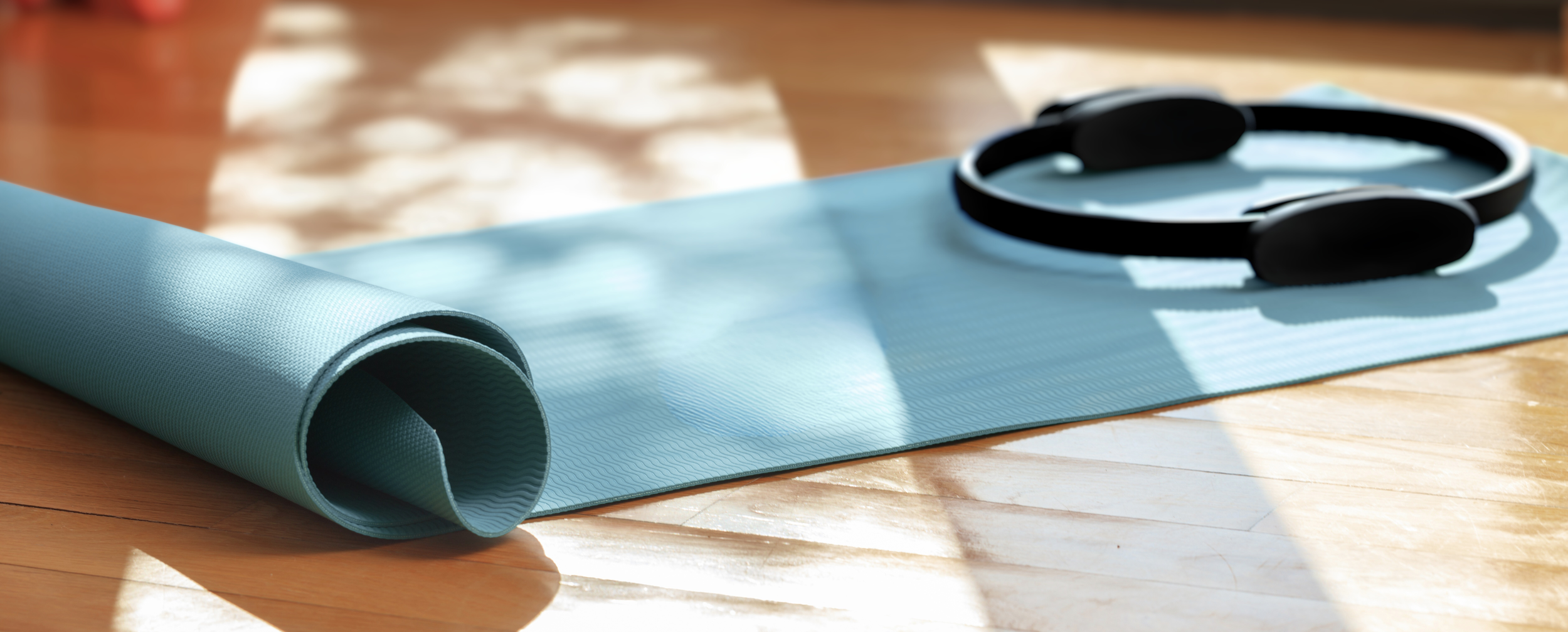 A light blue pilates mat is partially rolled on a wooden floor, with a black pilates magic ring resting on the unrolled portion.