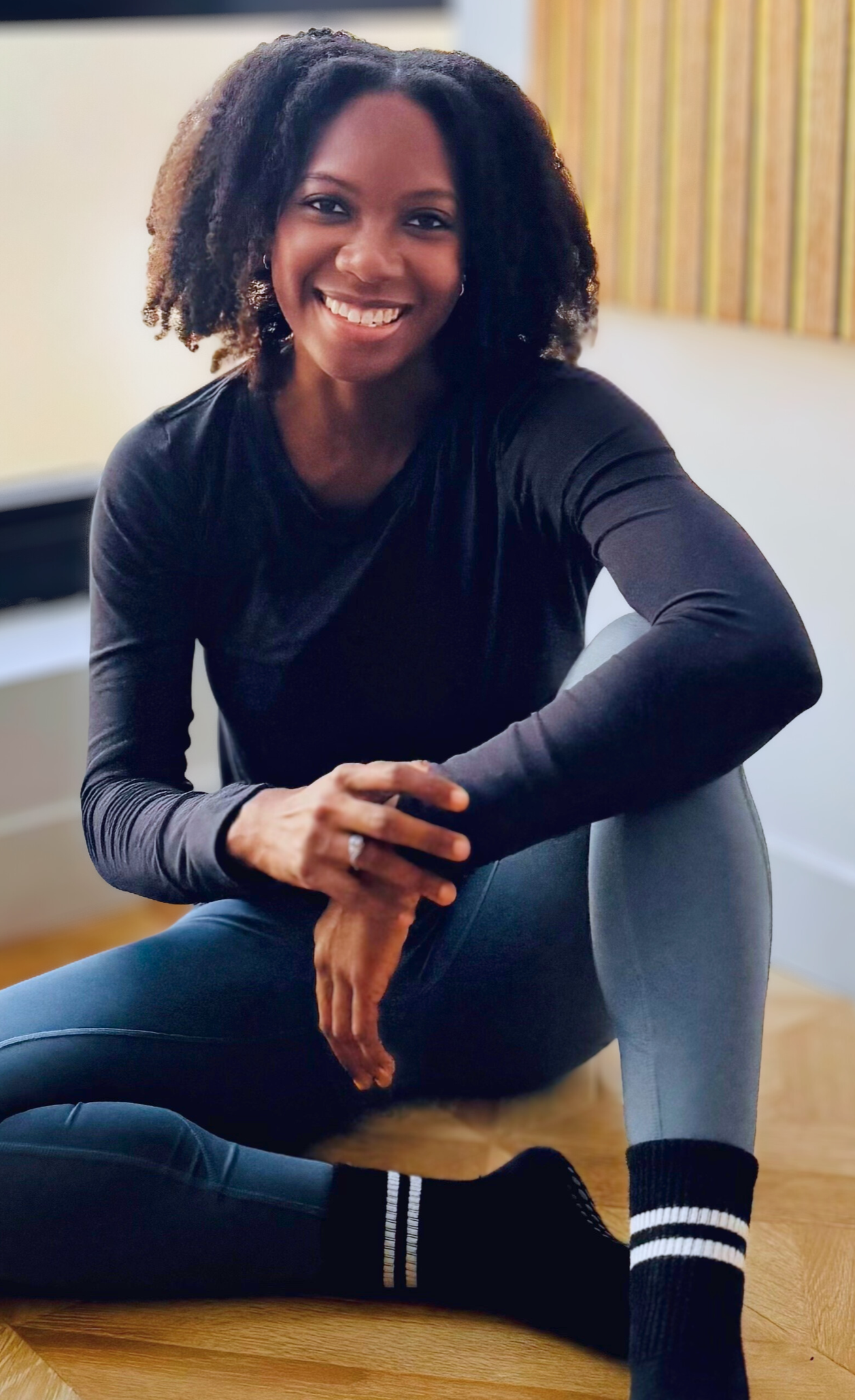 Courtney, a pilates apprentice and founder of True Align Pilates, sits on a wooden floor in a studio, smiling, and looking at the camera. She wears a dark long-sleeved shirt and grey leggings.