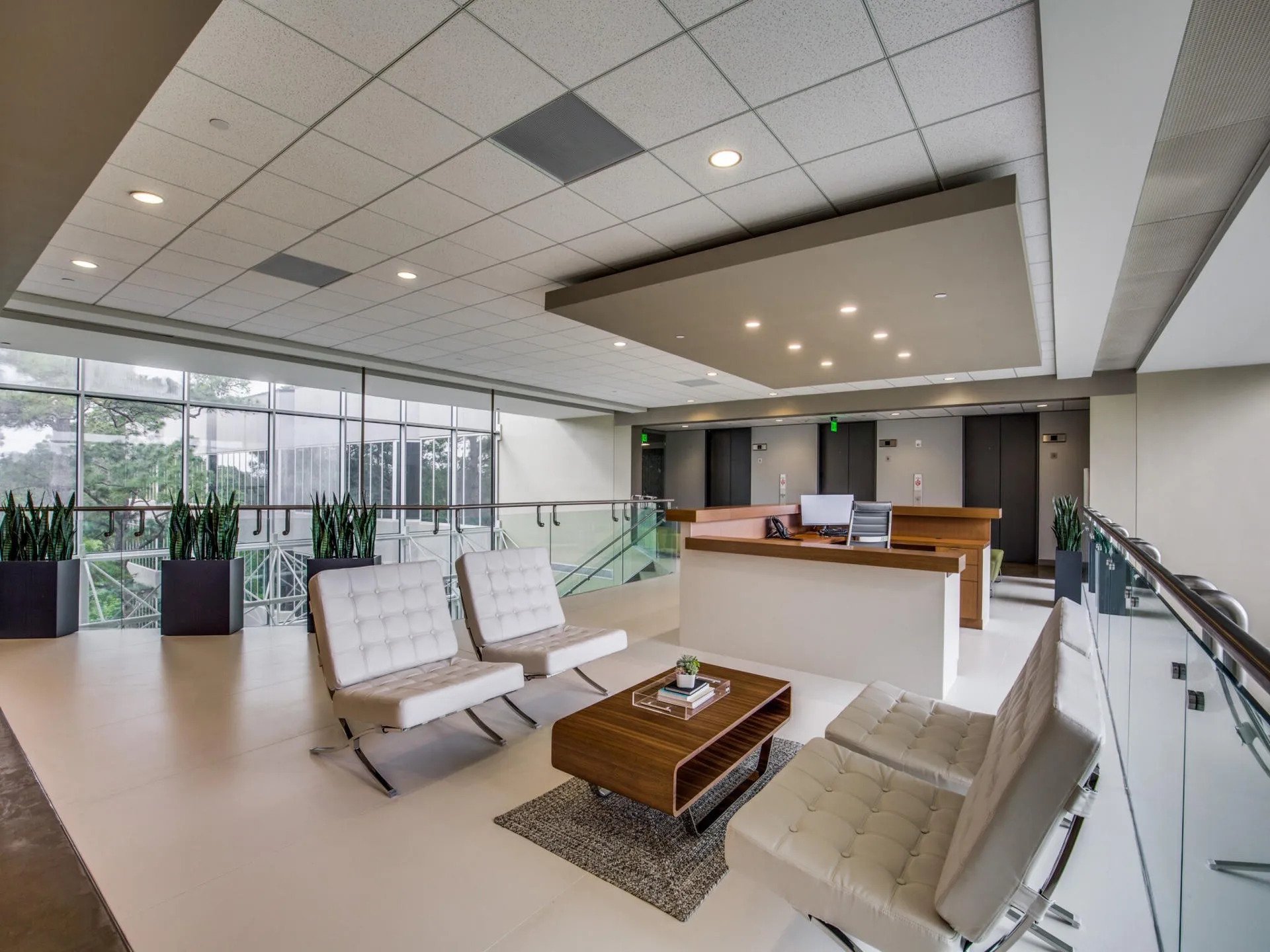 Modern office lobby with white leather chairs, a wooden coffee table, potted plants, and elevator doors in the background.