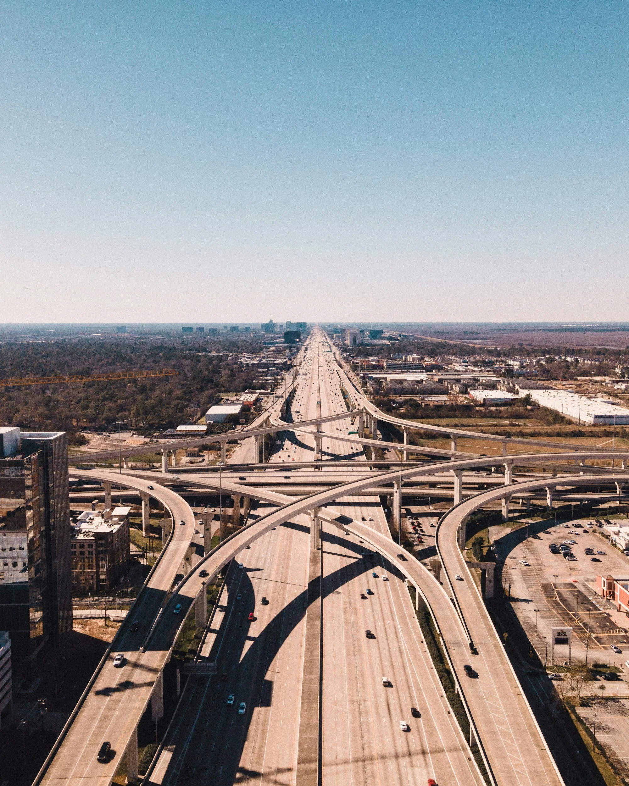 Aerial view of a large highway interchange with multiple overpasses and roads with moving cars in a cityscape with buildings and trees in the background.