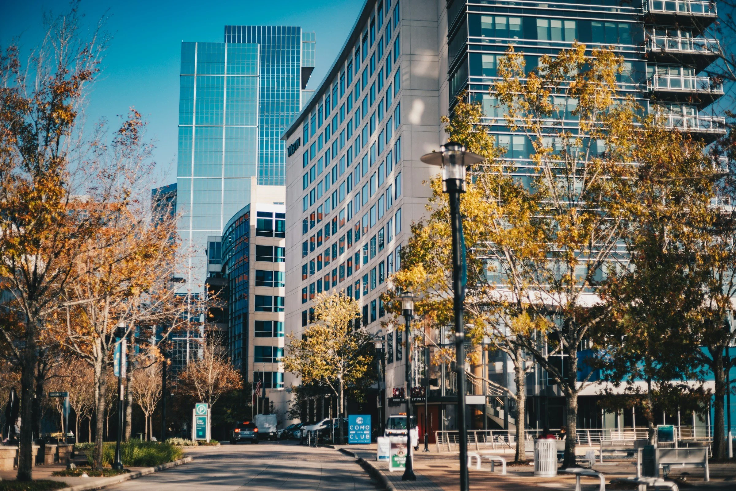 Urban scene with modern high-rise buildings, trees with fall foliage, street lamps, parked cars, and a clear blue sky.