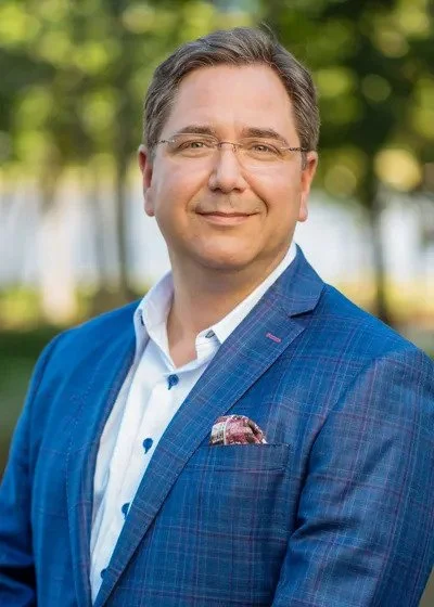 A man wearing glasses, a white shirt, and a blue suit jacket standing outdoors with greenery in the background