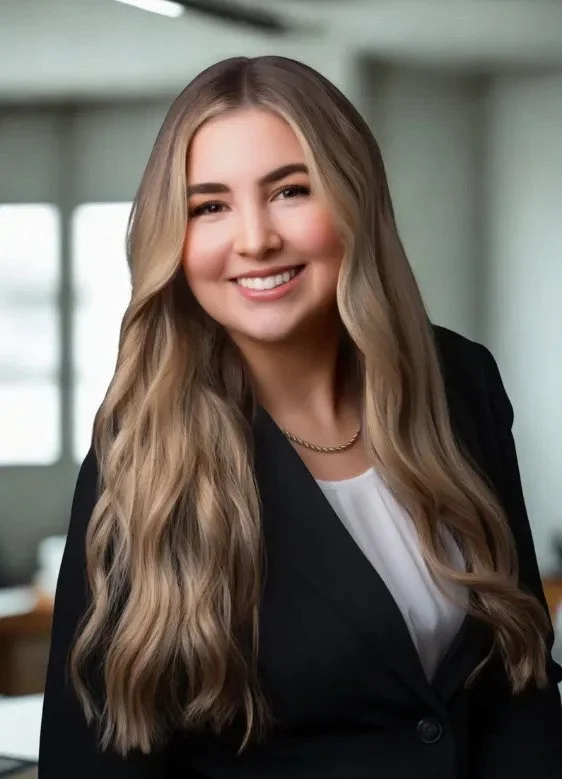 A young woman with long, wavy blonde hair, smiling, dressed in a black blazer and white top, standing in an office setting with windows in the background.
