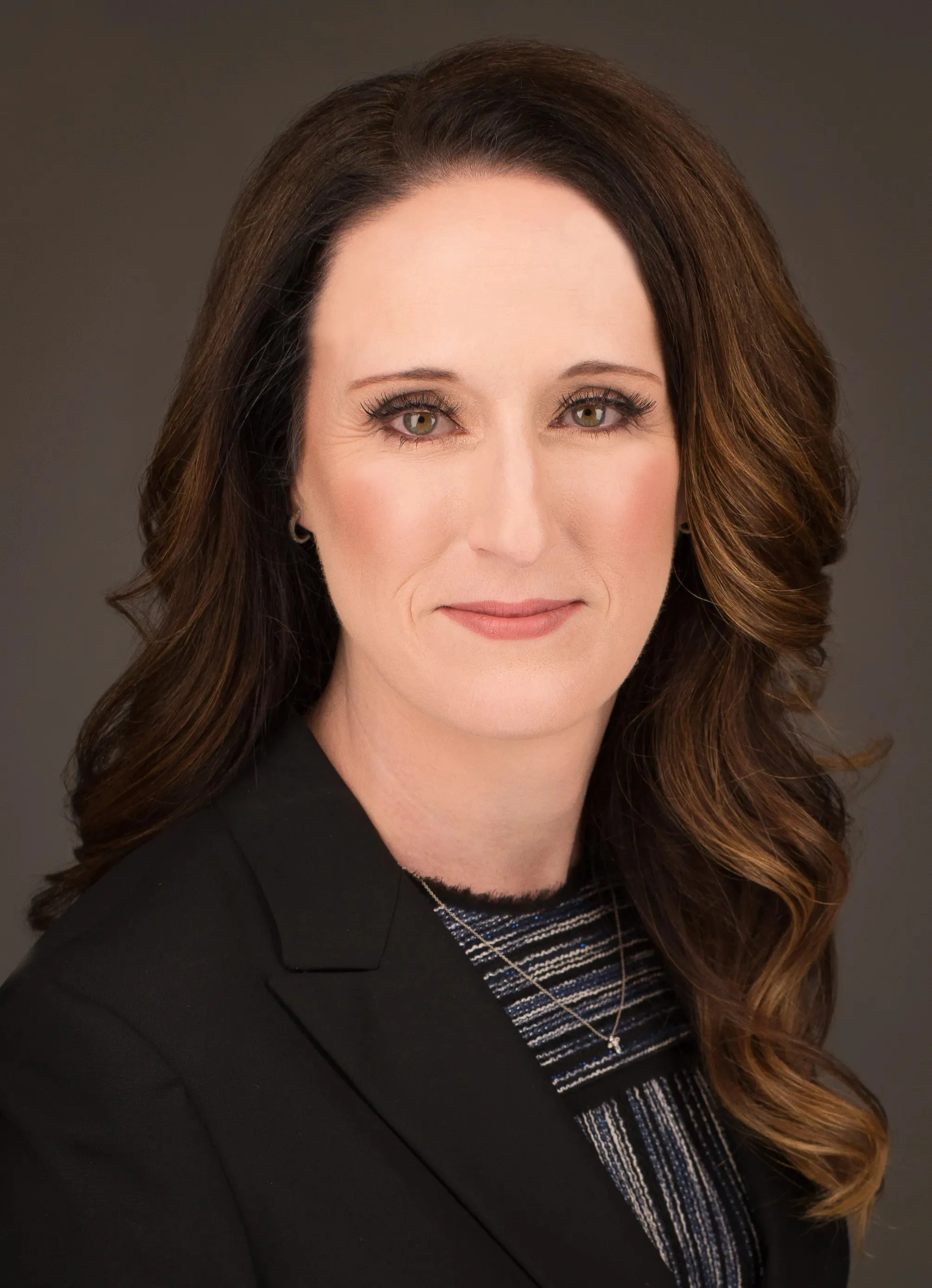 A professional woman with long, wavy brown hair and green eyes, wearing a black blazer and a striped top, against a dark gray background.