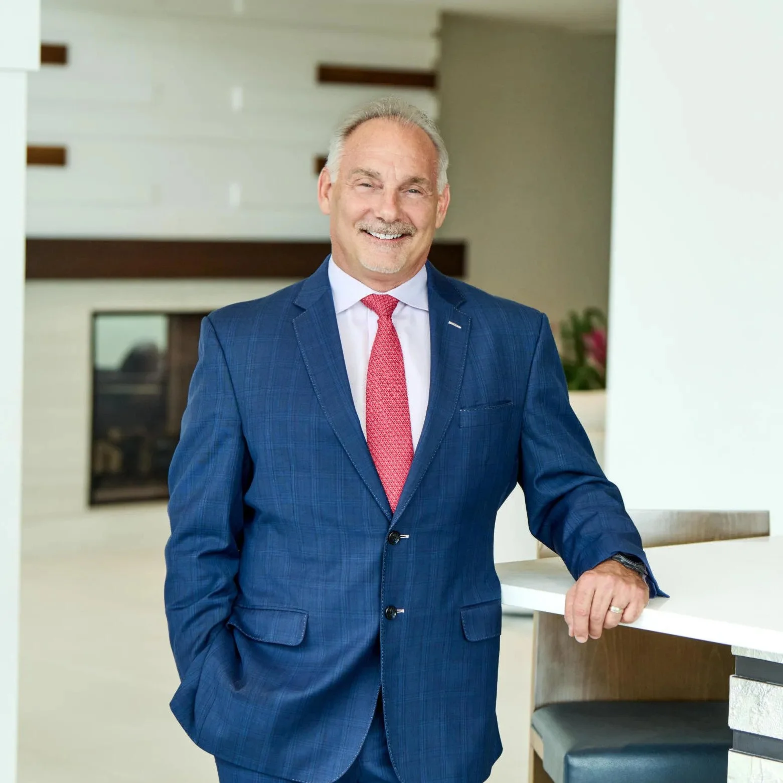 A professionally dressed man in a blue suit with a pink tie standing in a modern indoor setting, smiling.