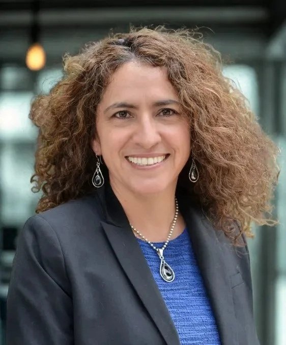 A woman with curly brown hair wearing a black blazer and blue top, smiling, with earrings and a necklace, standing indoors with large windows in the background.