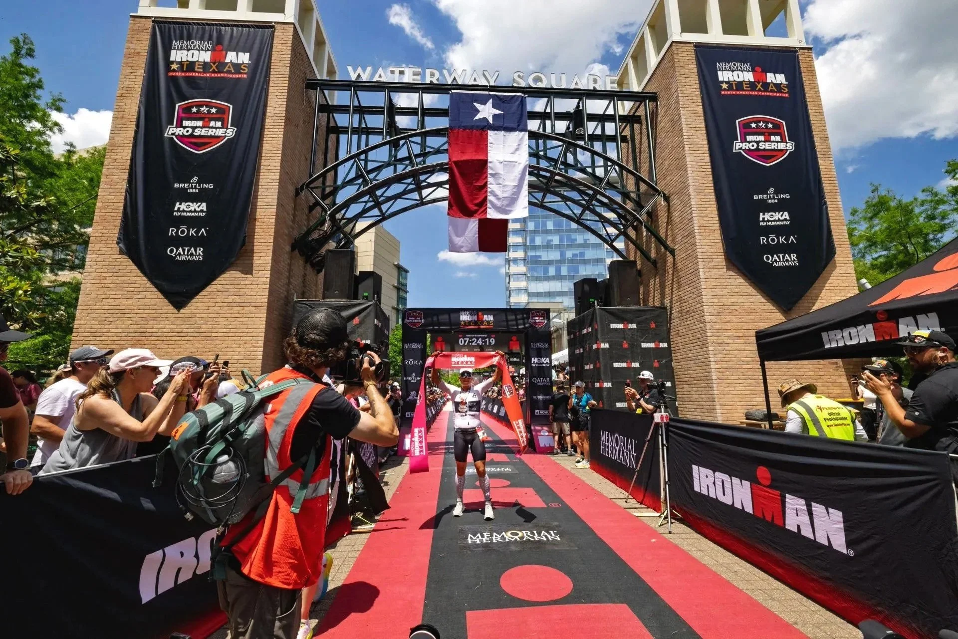 A person crossing the finish line at the Ironman Texas race, with arms raised in victory, surrounded by spectators and photographers, under banners and a Texas flag.