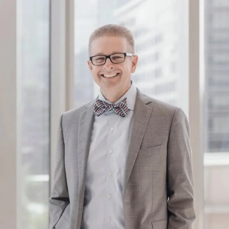 A smiling man wearing glasses, a gray suit, a white shirt, and a plaid bow tie, standing in front of a large window in a high-rise building.