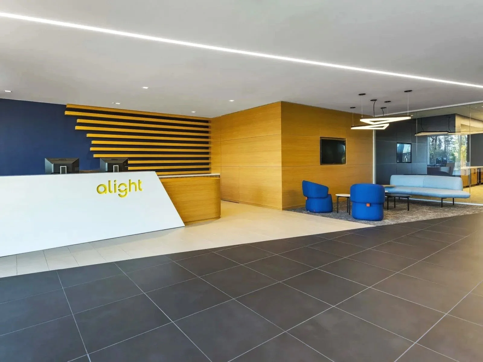 Modern hotel lobby with a reception desk labeled 'alight', blue and white seating area, wooden accents, two wall-mounted televisions, black tile flooring, and large glass windows showing outdoor greenery.
