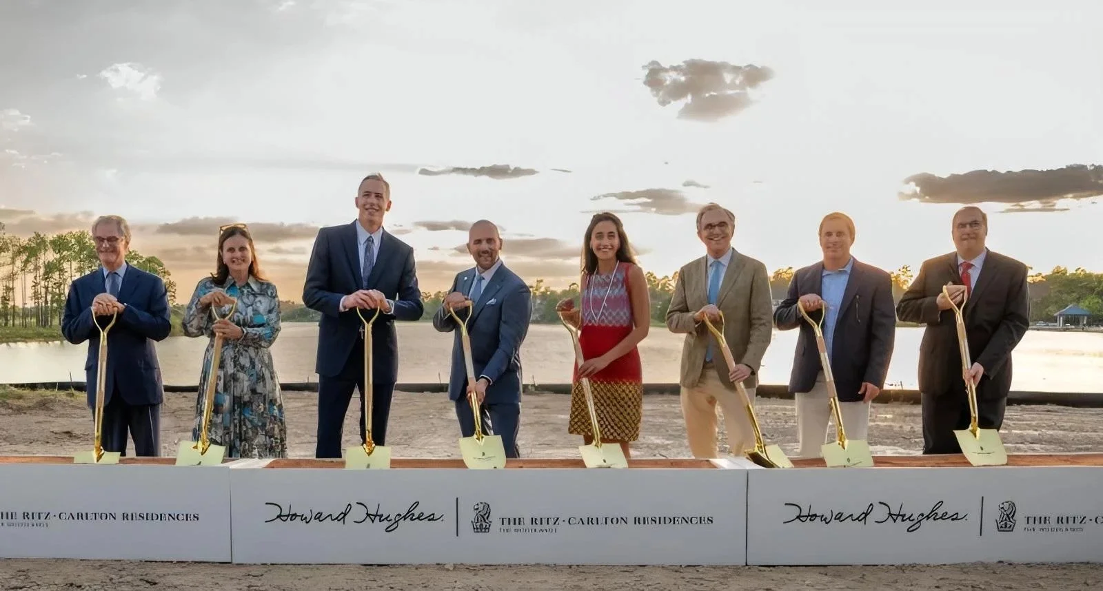 A group of nine people in formal attire holding golden-colored shovels at a groundbreaking ceremony near a body of water during sunset, with a sign reading "Howard Hughes."