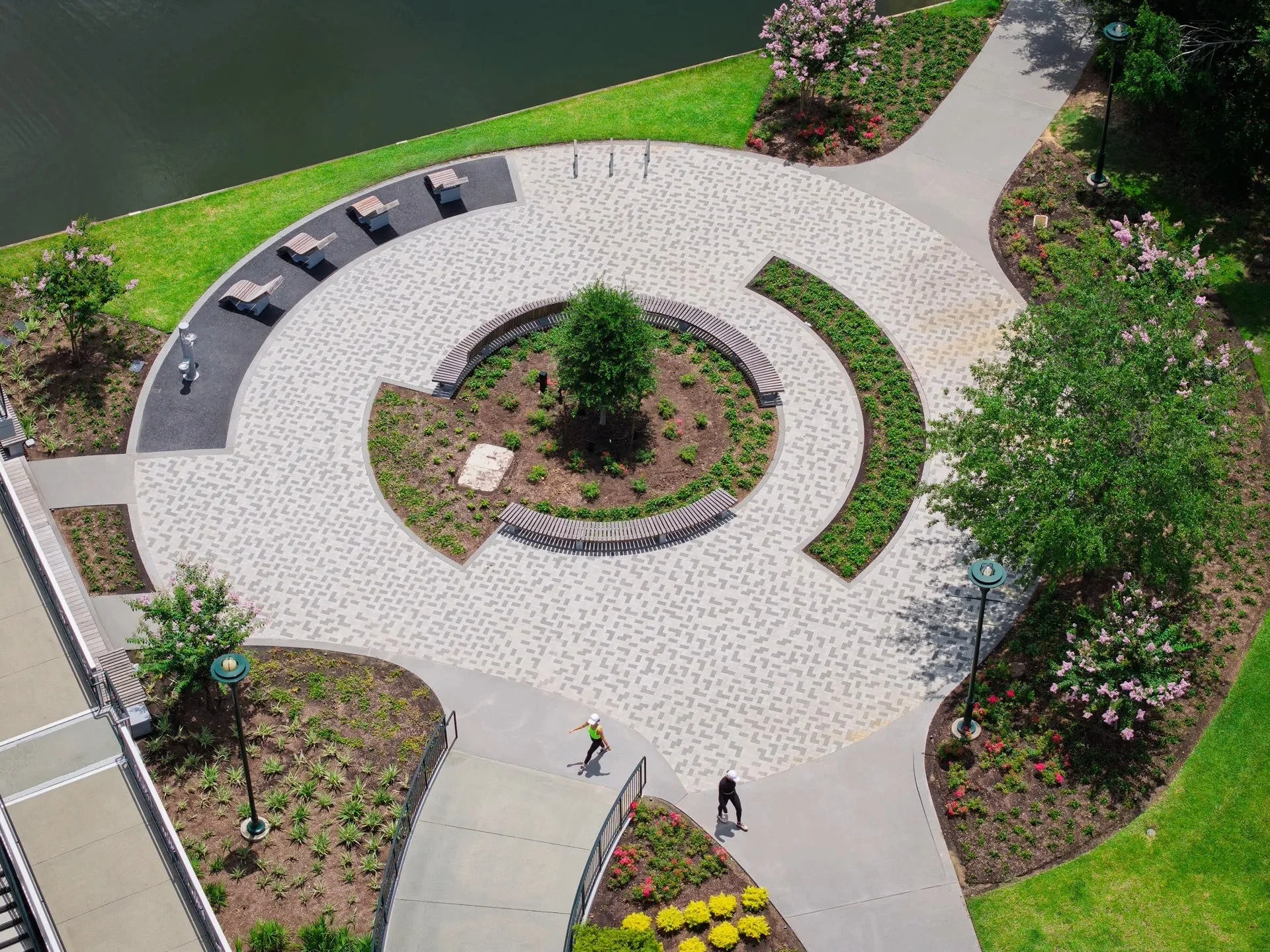 Aerial view of a park with a circular paved walkway around a central garden with a tree. There are benches, flower beds, trees, and walking paths surrounding the paved area.
