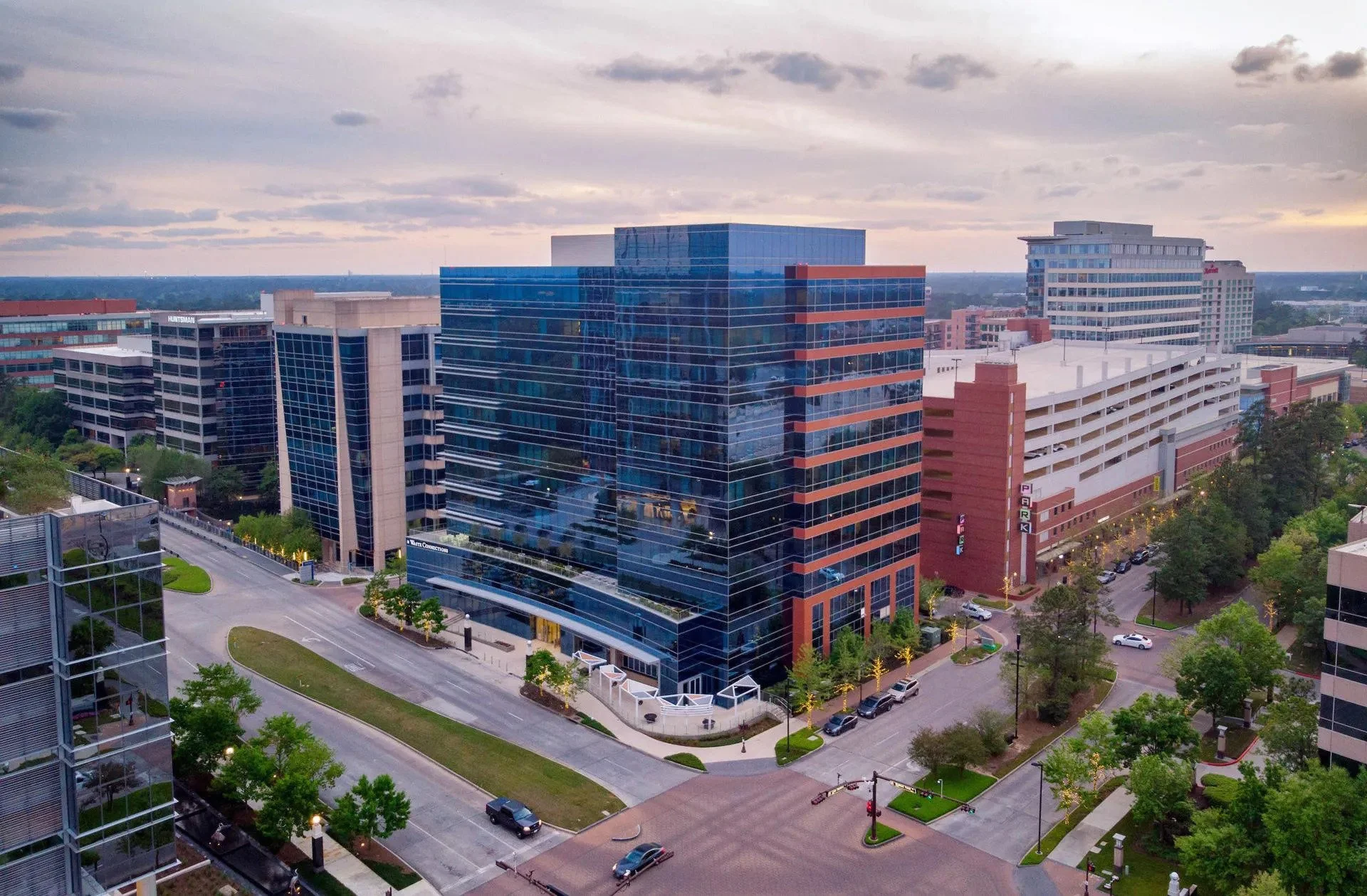Aerial view of modern office buildings with glass facades in a city during dusk, with streets, trees, and parked cars visible.