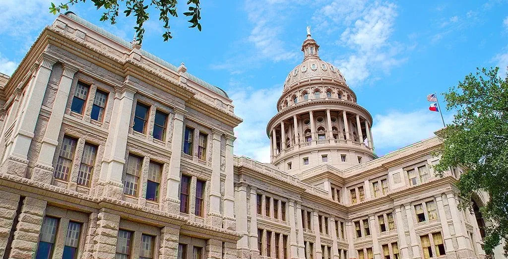 View of the Texas State Capitol building with a cloudy sky and green trees in the foreground.