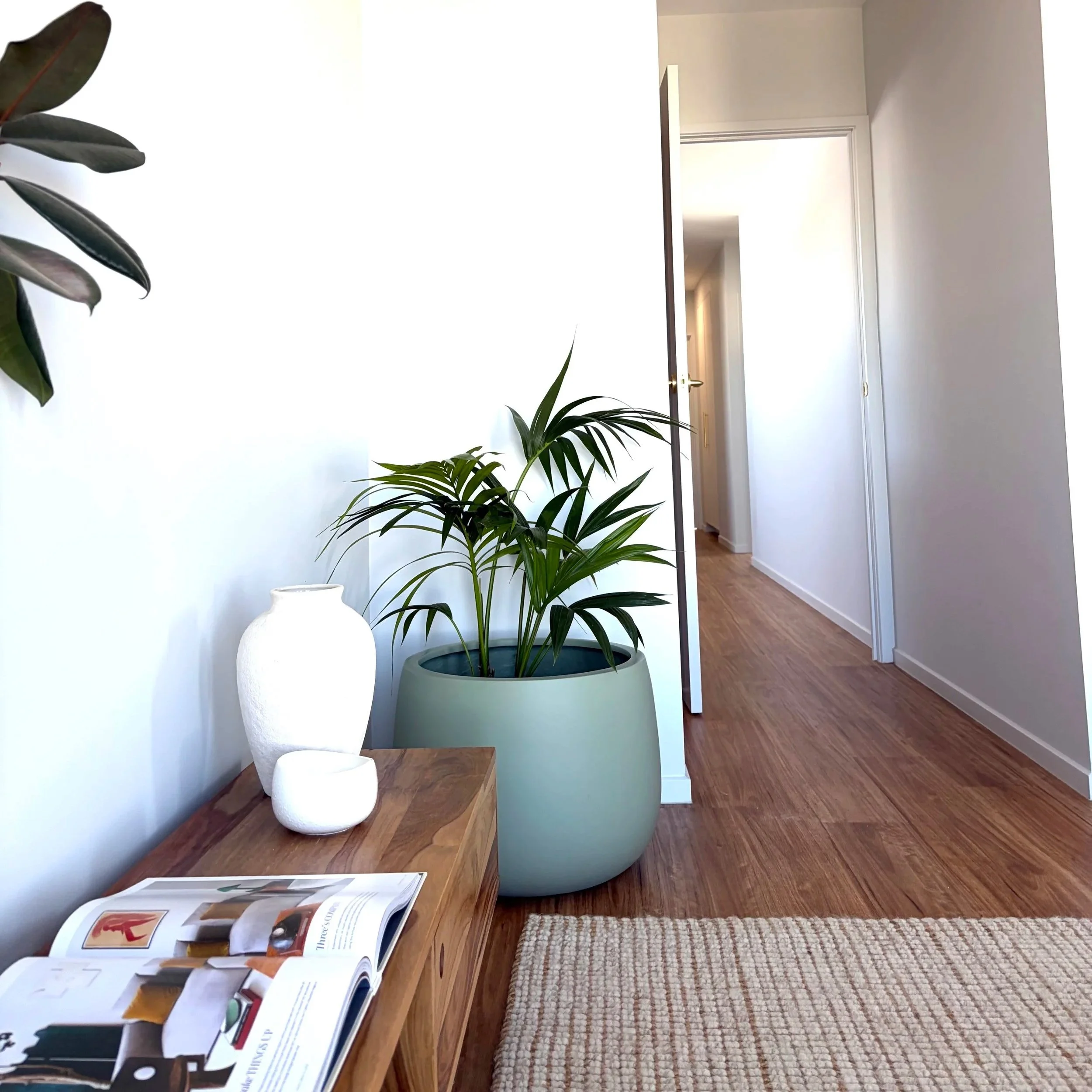 Interior scene featuring a wooden table with open magazines and newspapers, a white ceramic vase, a potted plant with long green leaves, a woven rug, and an open door leading to a hallway with white walls and wooden flooring.