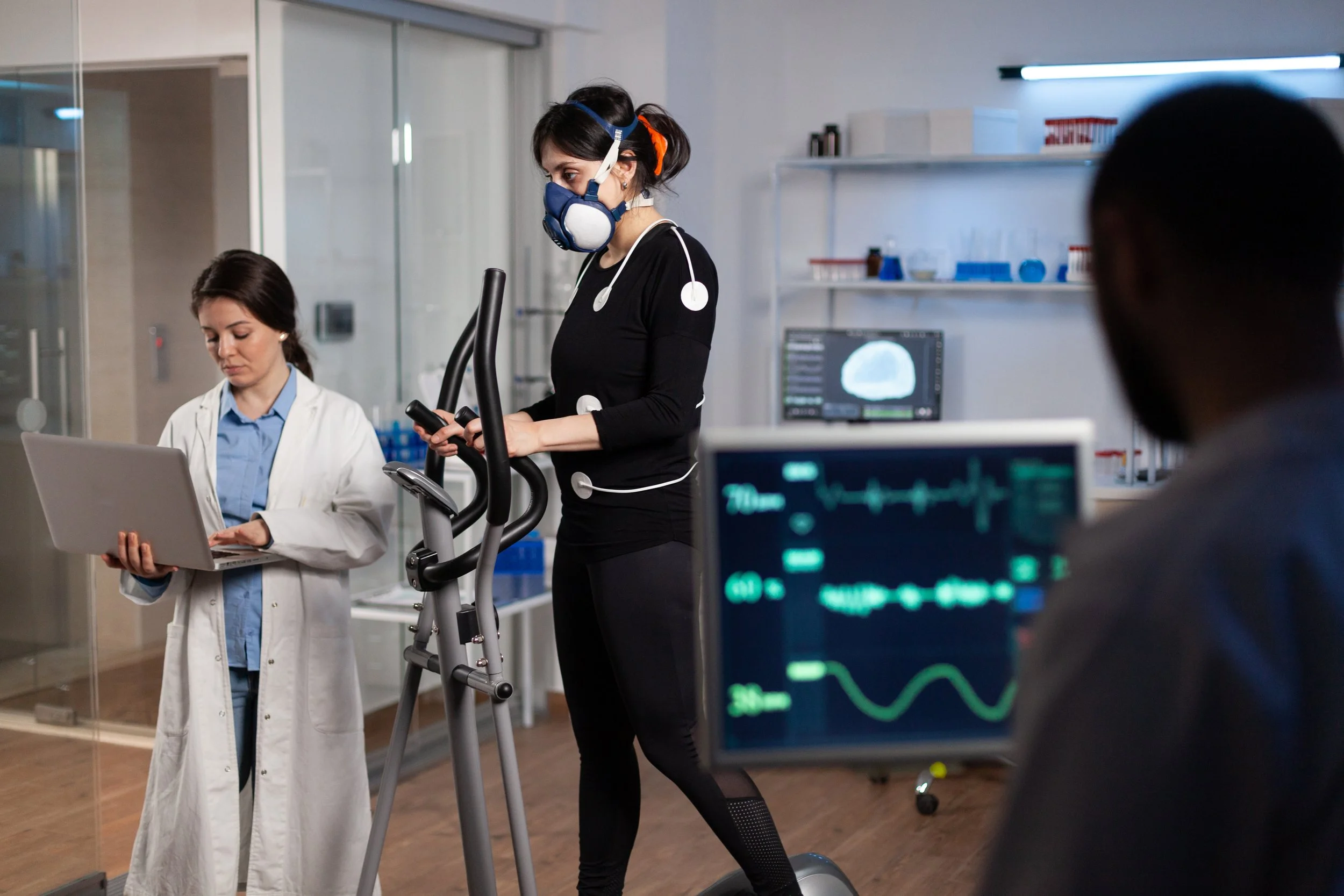 Medical professionals conducting a fitness test. One woman wearing a mask and fitness tracker on an exercise machine, another woman in a lab coat using a laptop, and a person with medical monitors in the foreground.