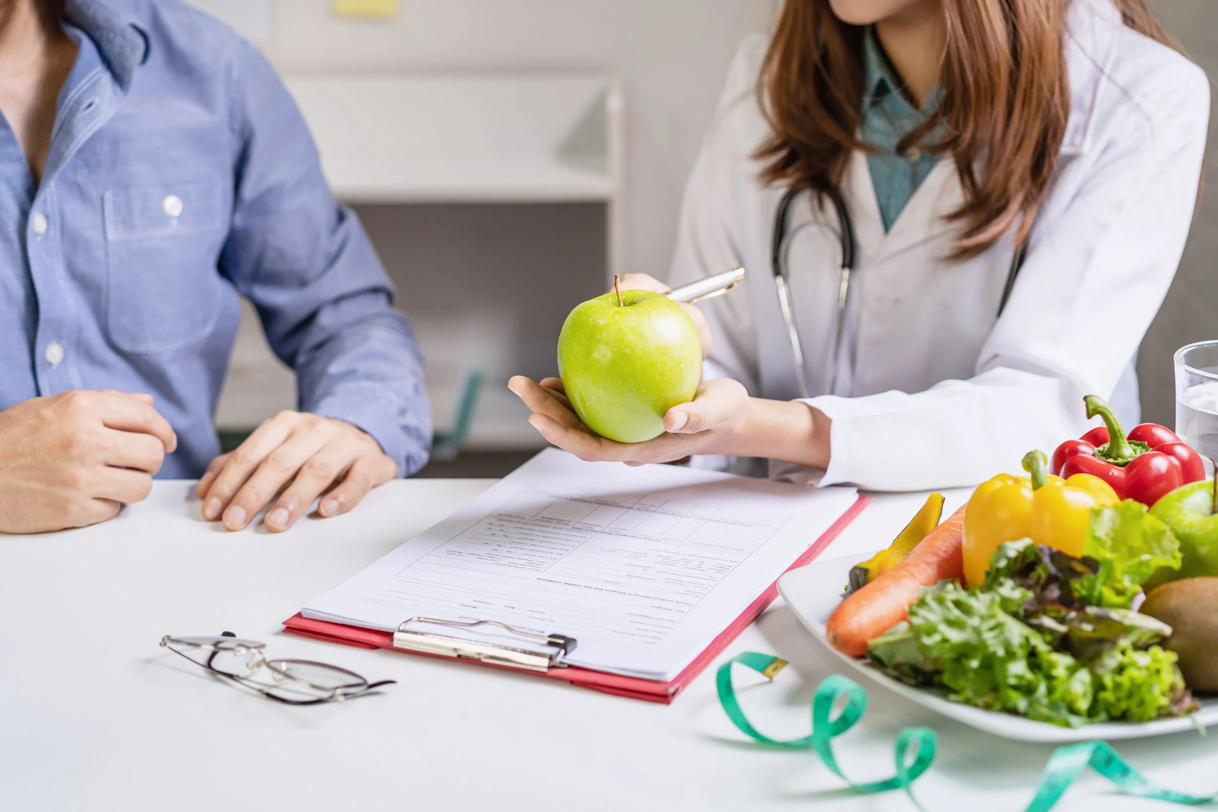 A healthcare professional showing a green apple to a patient, with a clipboard, glasses, and a bowl of vegetables on the table.