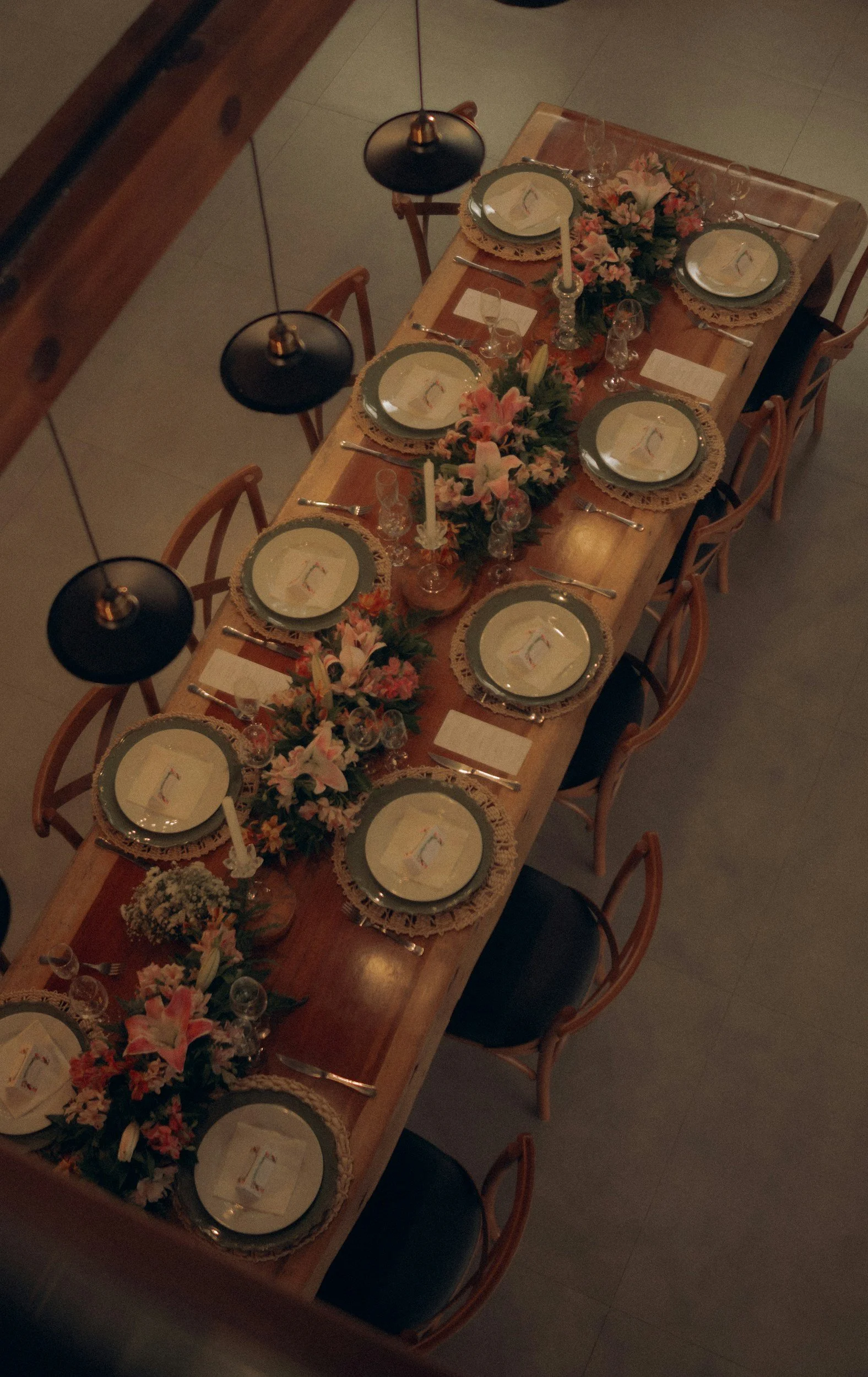 A long wooden dining table set for six, decorated with floral centerpieces, plates with lace doilies and small flags, glasses, cutlery, and candles surrounded by wooden chairs, viewed from above in a dimly lit room.