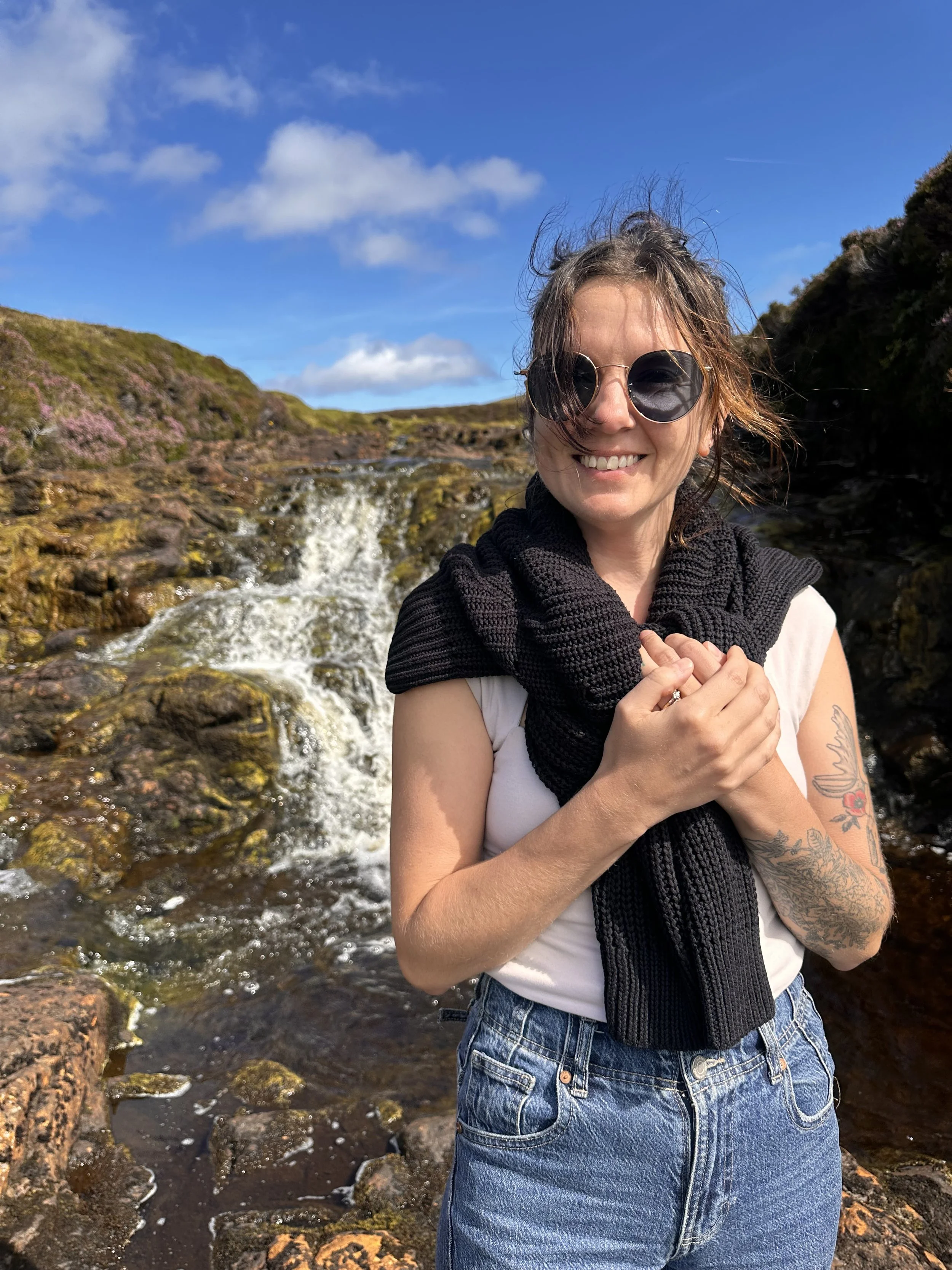 A smiling woman wearing sunglasses and a black scarf around her neck standing in front of a small waterfall in a rocky outdoor setting under a blue sky with scattered clouds.
