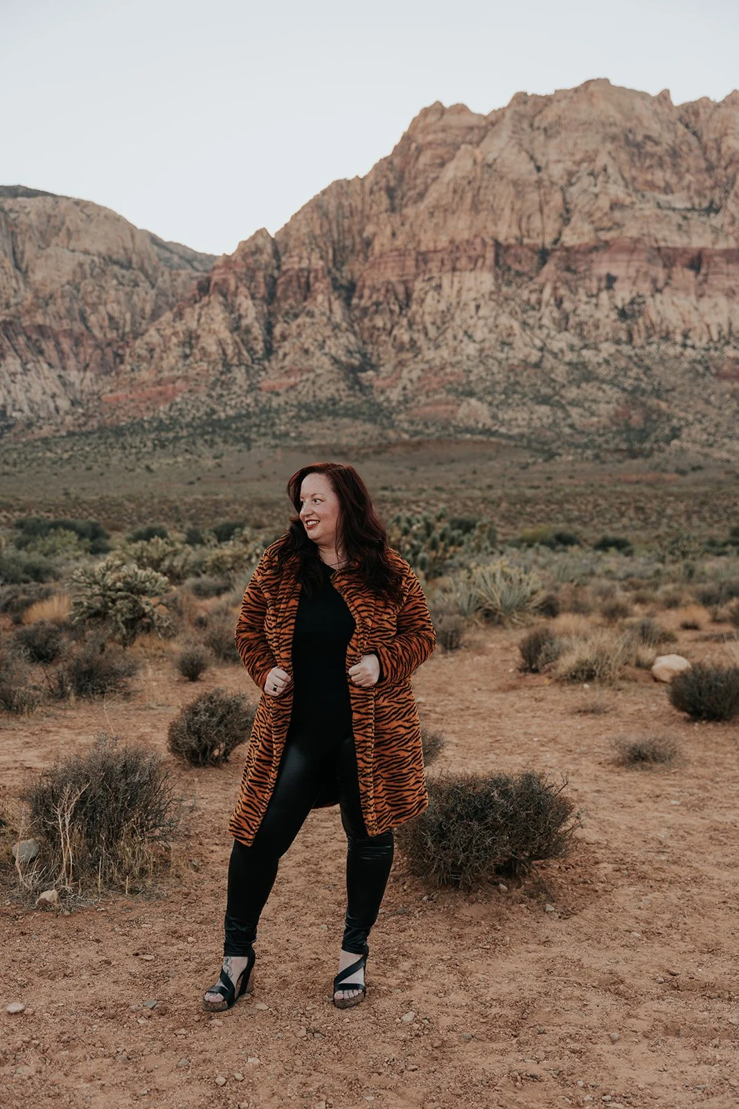 Woman standing in her power in tiger-striped coat and black outfit standing in a desert landscape with mountains in the background.