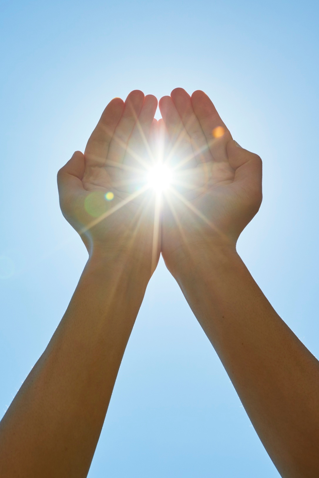 Hands clasped together with the sun shining through the gap, against a clear blue sky.