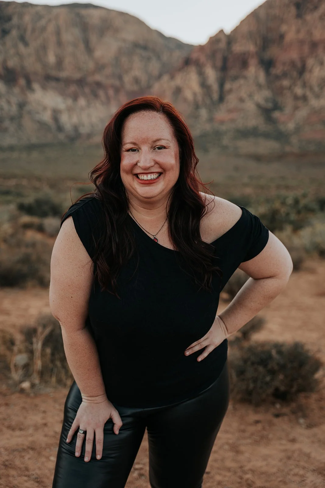 A self regulated woman with long dark hair smiling and standing outdoors in front of mountains with a desert landscape, wearing a black top and black pants.