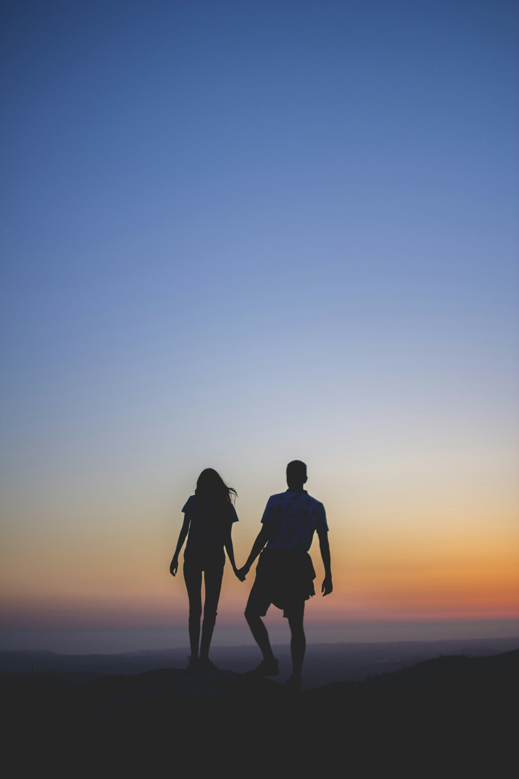 Silhouette of a couple holding hands on a hilltop during sunset, with a gradient sky transitioning from orange to blue.