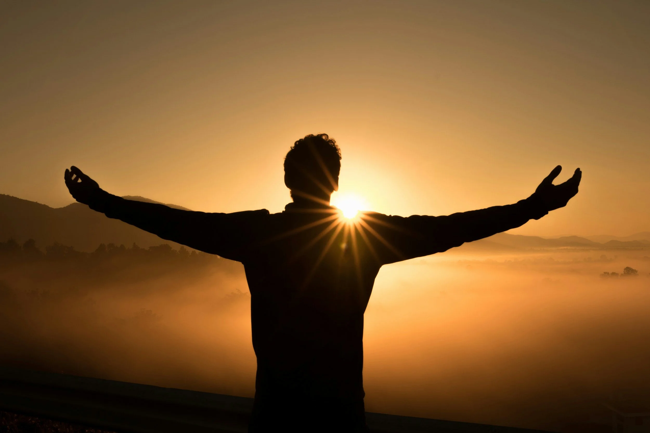 A person with outstretched arms standing outdoors at sunrise or sunset, with the sun positioned behind their head, creating a silhouette effect and a starburst of light.