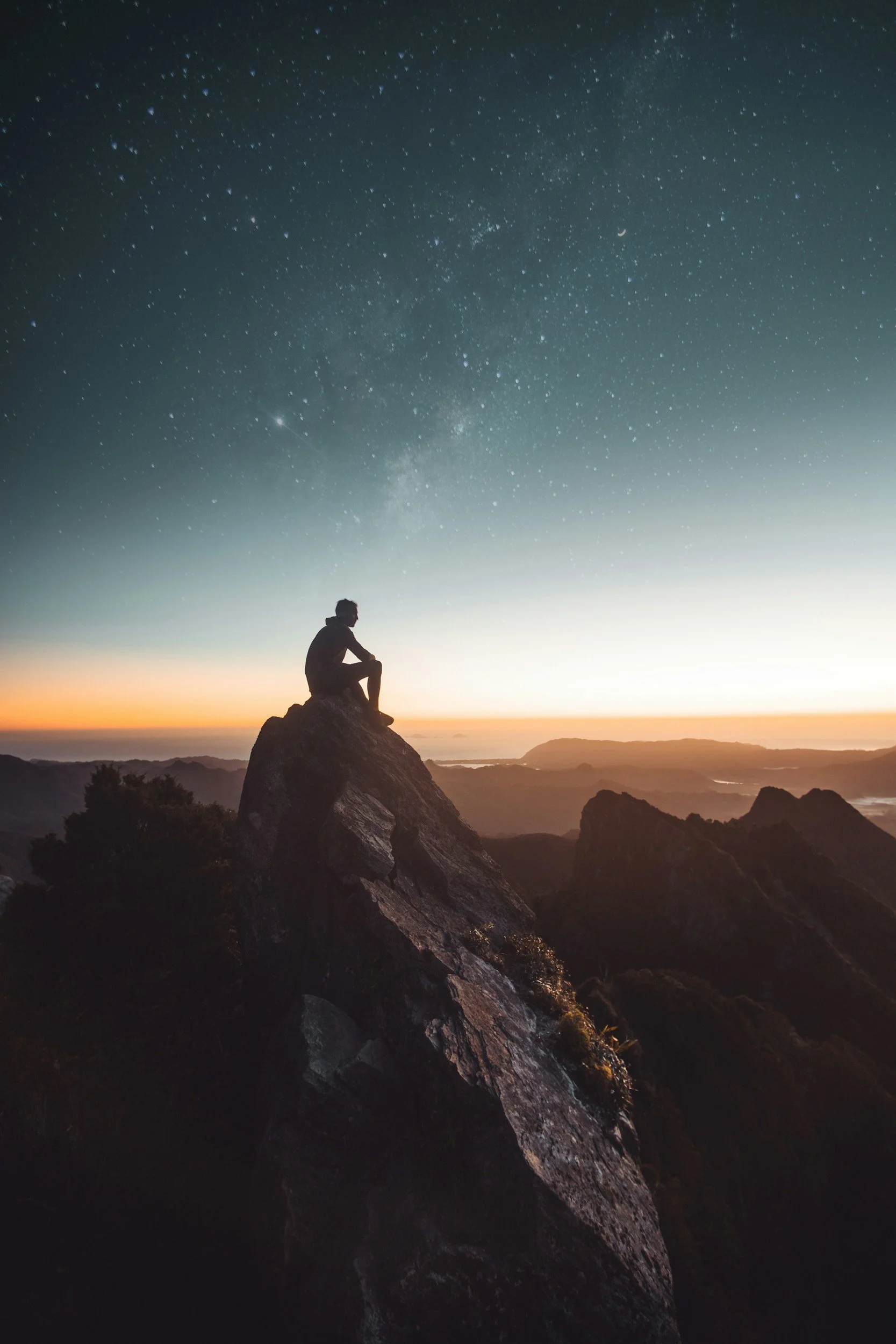 A person sitting on a large rock formation during dusk with a starry night sky, including the Milky Way galaxy, in the background.