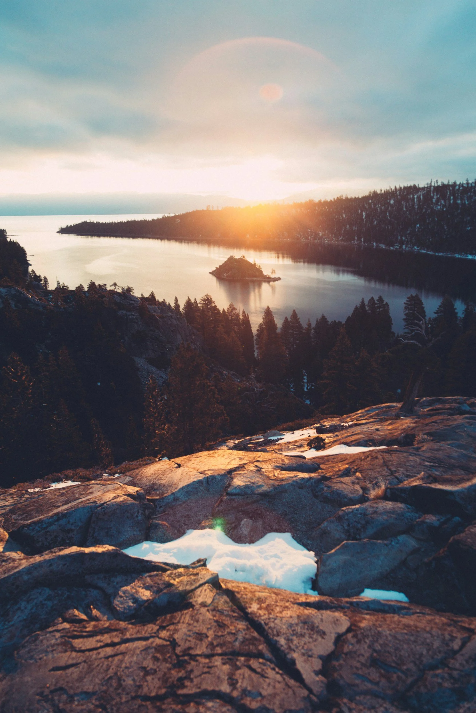 Sunset over a lake with a small island and forested hills, viewed from a rocky overlook with patches of snow.