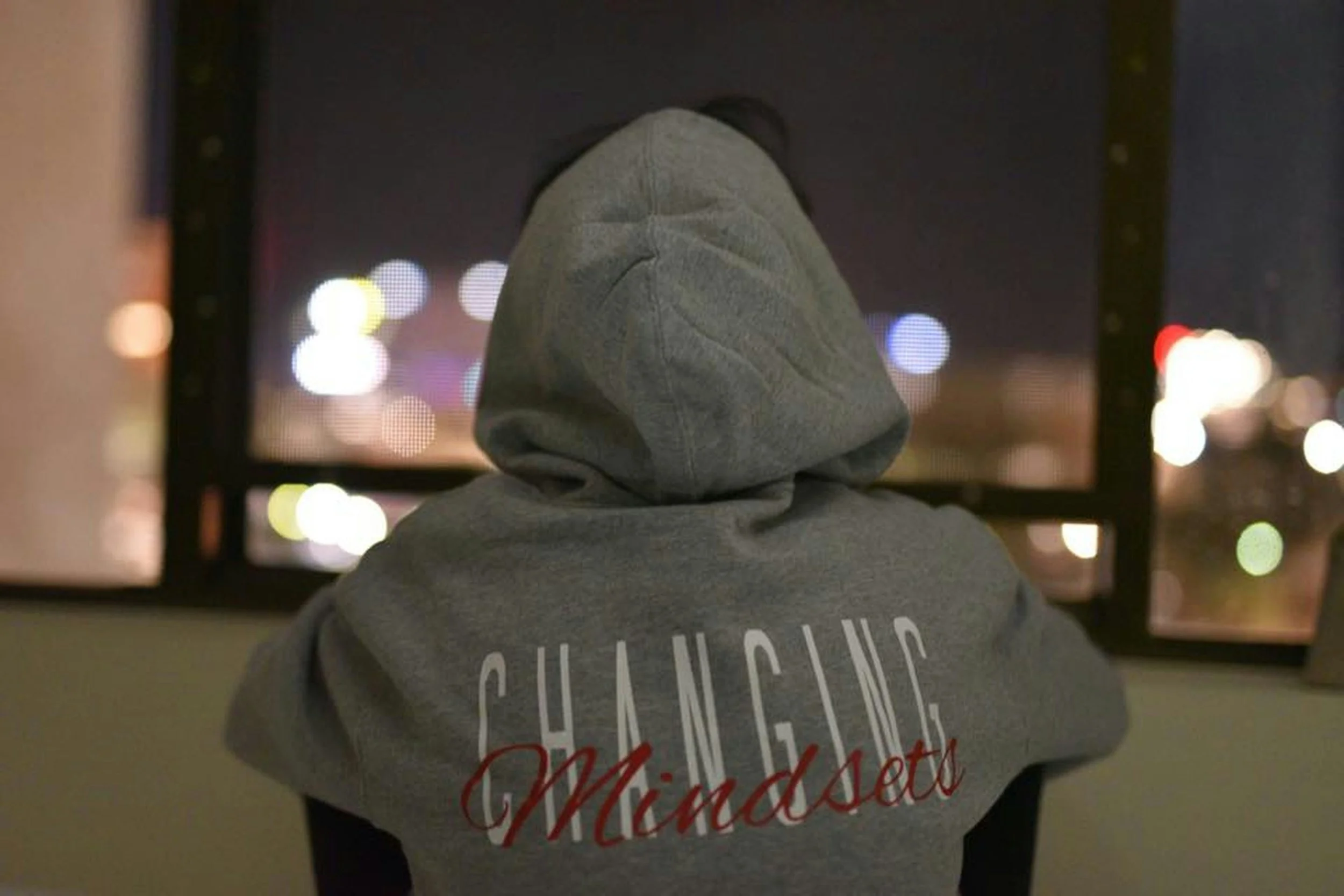 A person in a gray hoodie with the words 'CHANGE MINDSET' printed on the back, looking out at a window at night with blurred city lights in the background.