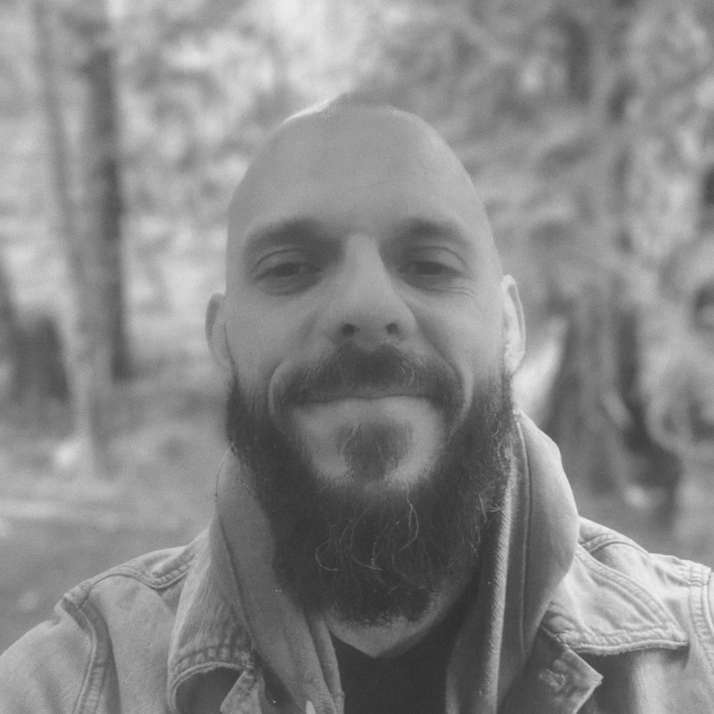 Black and white close-up selfie of a man with a beard and mustache, smiling slightly, outdoors with blurred trees in the background.