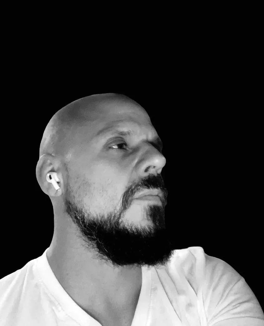 Black and white photo of a man with a beard, wearing a white shirt, looking to the right, with earbuds in his ears, against a solid black background.