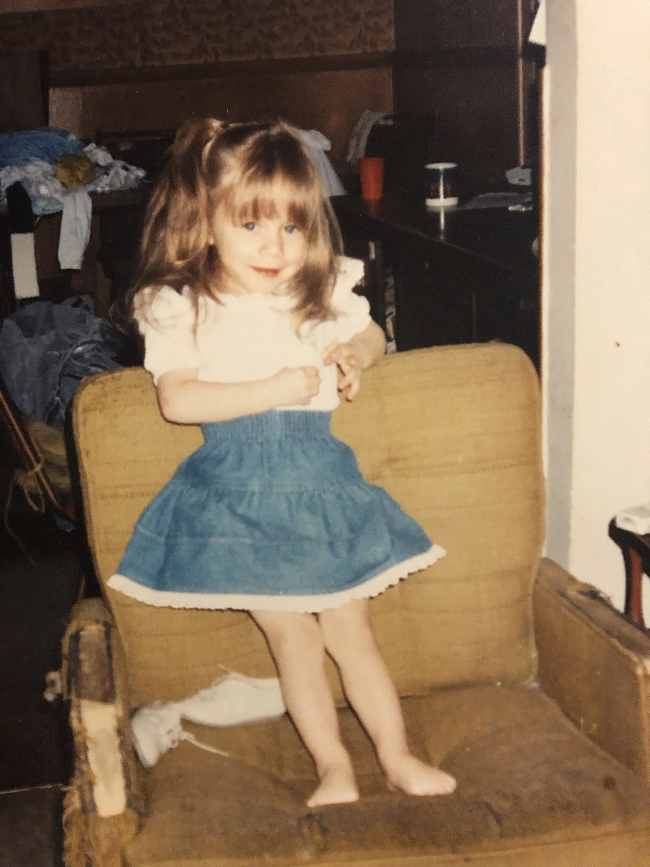A young girl (Ashley) with wavy brown hair in a white top and blue skirt, standing with her hands near her chest in a room with wooden furniture and cluttered laundry or clothes around. She has a slight smile on her face.