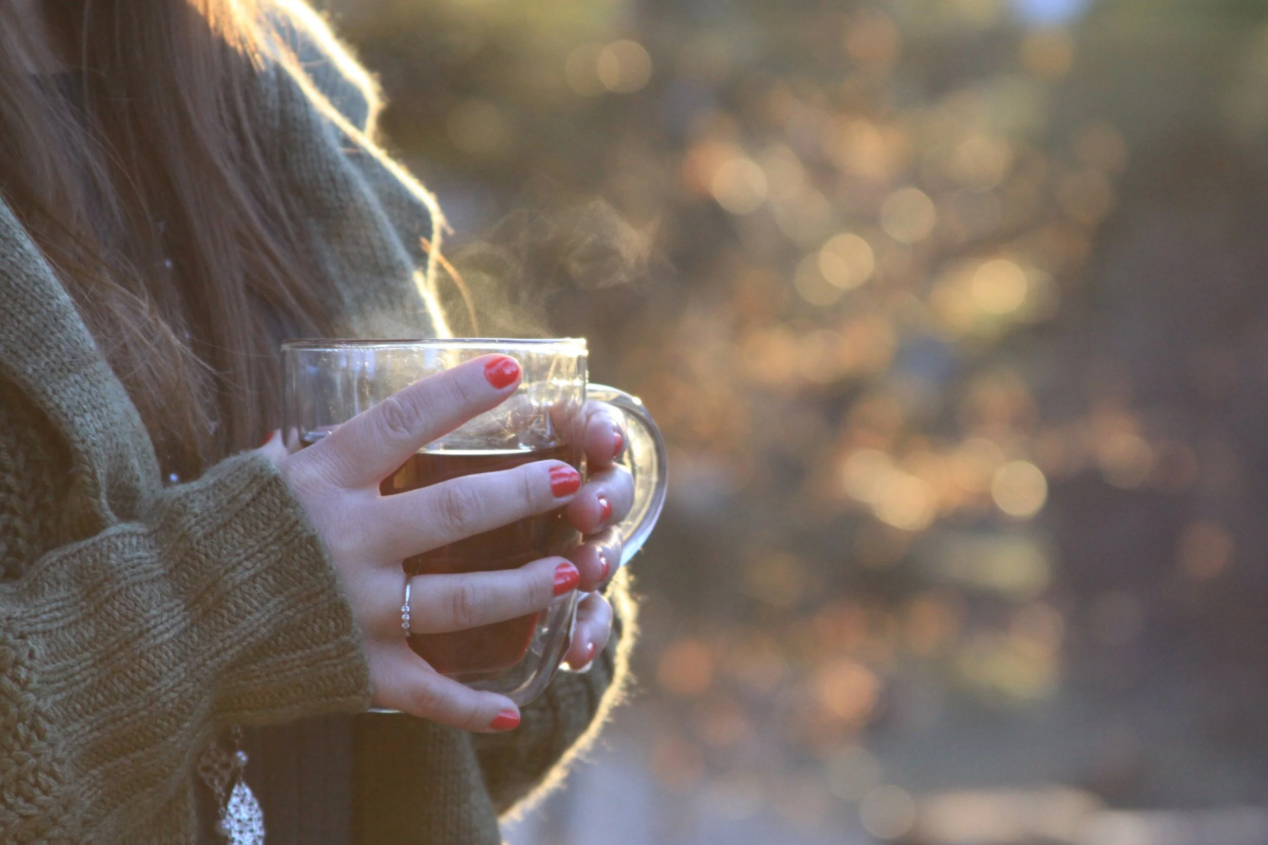 Person holding a glass mug with a hot beverage, wearing a brown knit sweater, with their nails painted red, outdoors during golden hour.