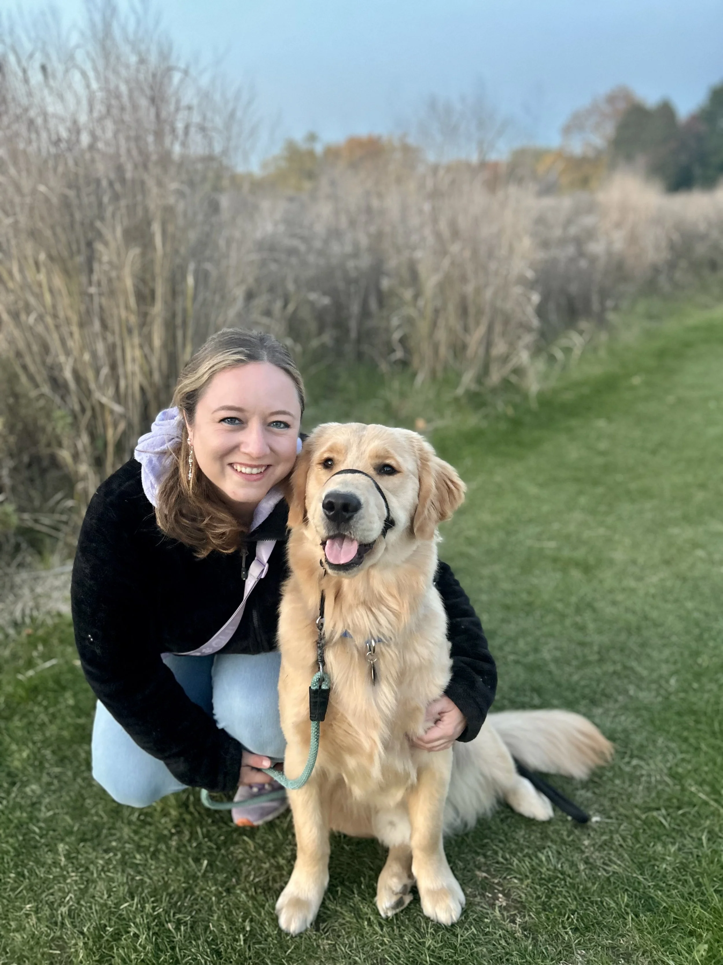 A woman (Ashley) smiling while hugging a golden retriever dog outdoors on a grassy area, with trees and a clear sky in the background.