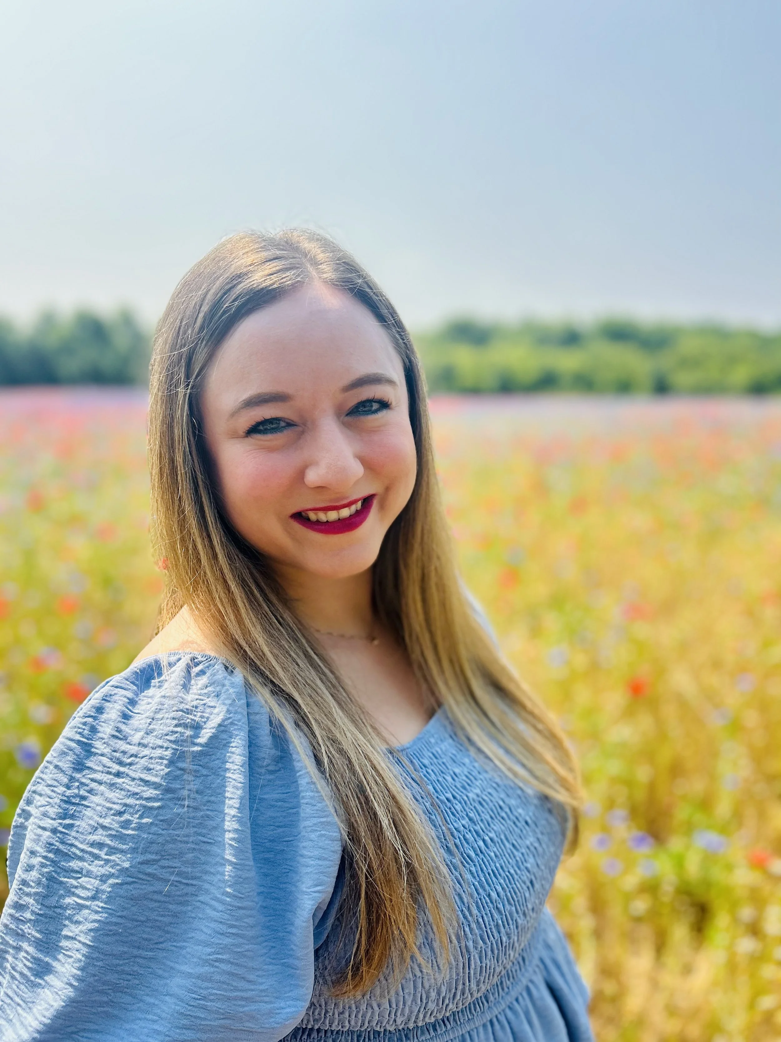 A woman (Ashley) with long blonde hair and bright red lipstick smiling outdoors in a field of colorful flowers, wearing a light blue dress.