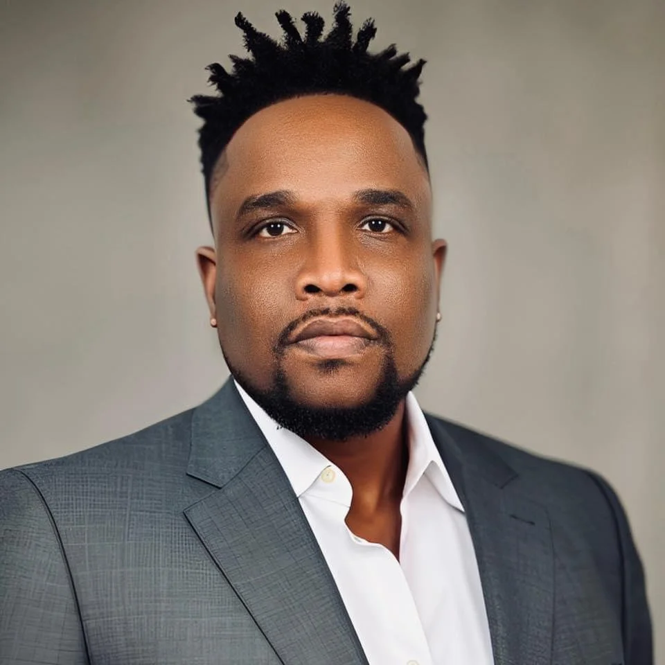 A portrait of a man in a suit with a serious expression, dark hair styled in twists, and a beard, against a plain background.