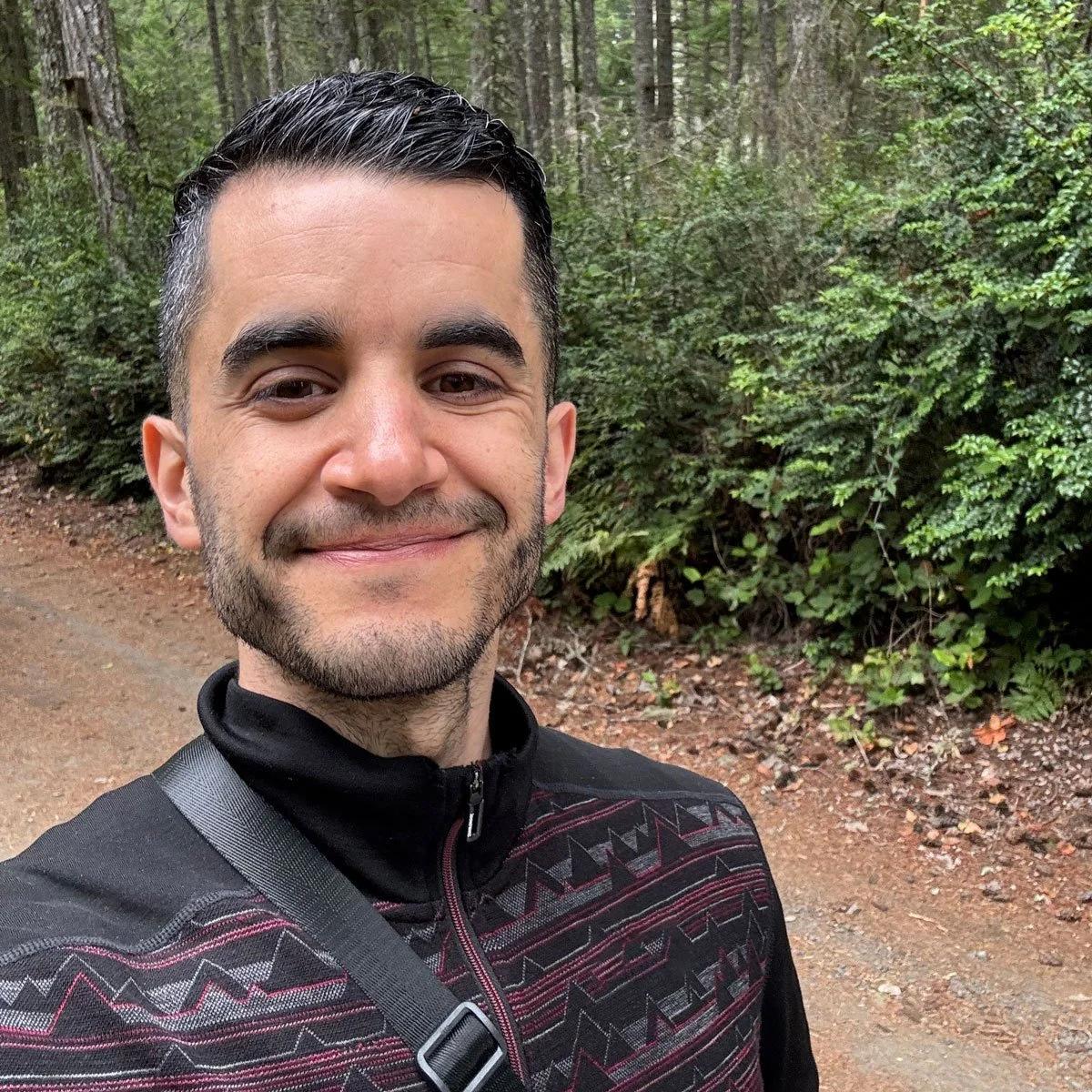 A smiling man with dark hair and a beard taking a selfie outdoors on a forest trail.
