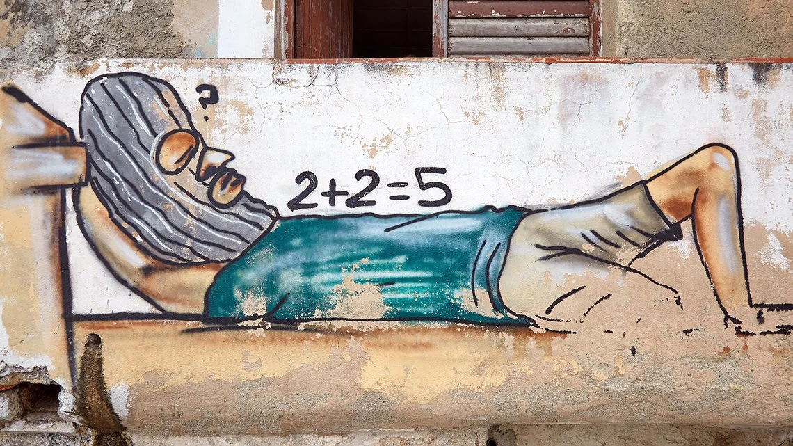 A street mural of a man lying on his side, wearing glasses and a striped cap, with a beard. The man is depicted in a relaxed position with his hand under his head. A math equation '2+2=5' is painted next to him.
