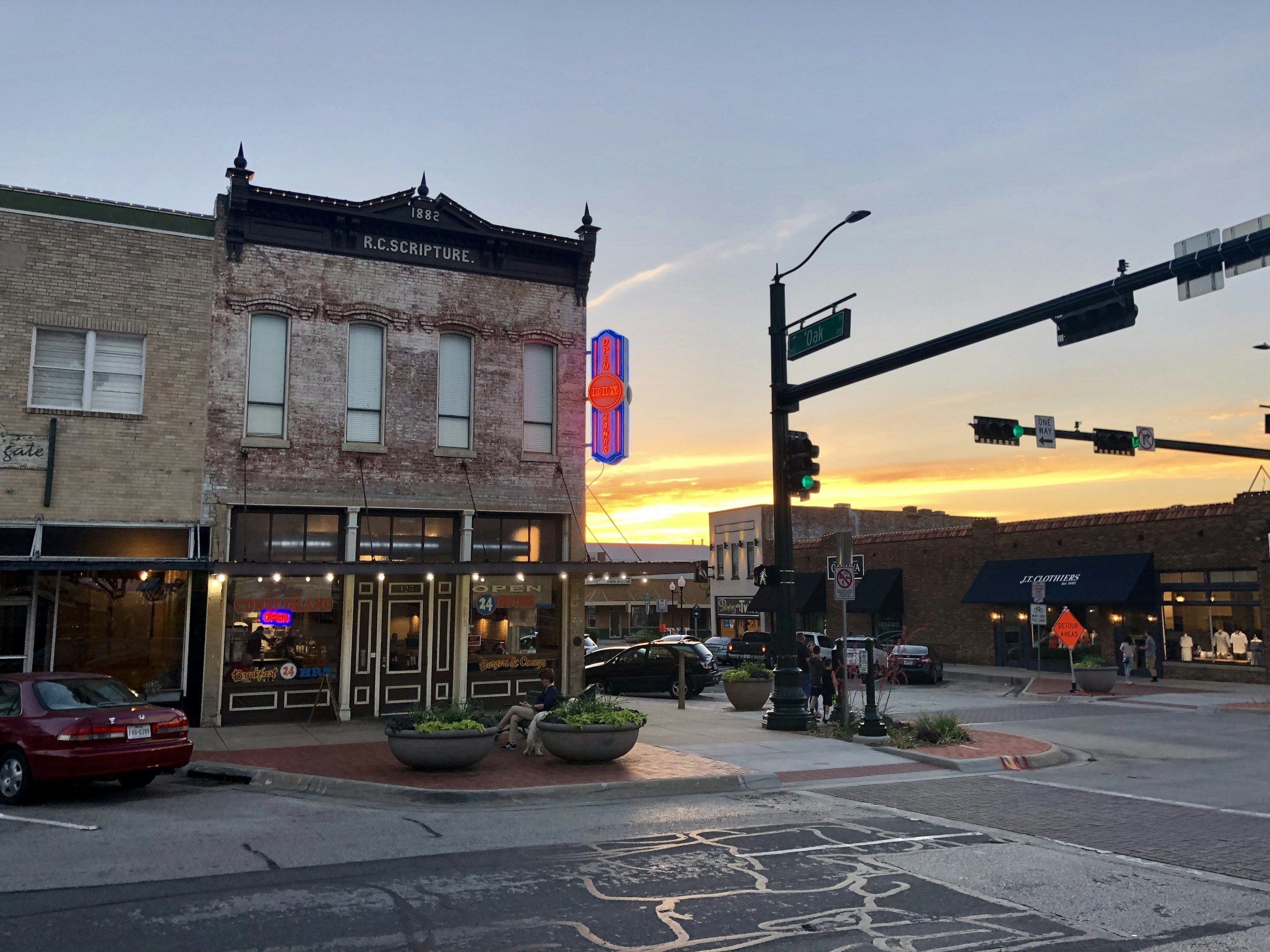 Street view at sunset with historic building, a neon sign, cafe, and pedestrians in a downtown area.