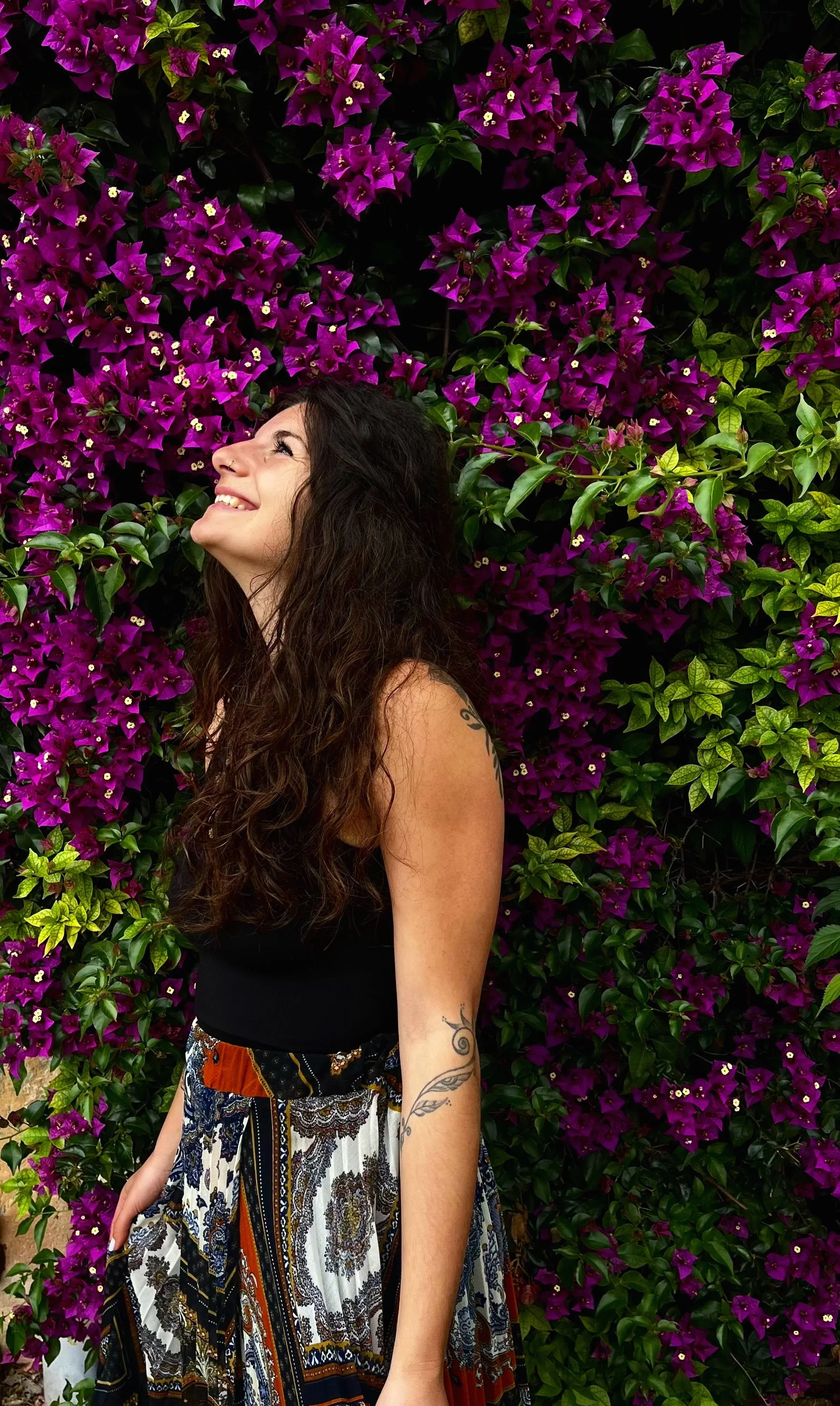Frau mit langen, lockigen Haaren und Tätowierungen an den Armen, die vor einer Wand aus magentafarbenen Bougainvillea-Blüten steht und lächelnd nach oben schaut.