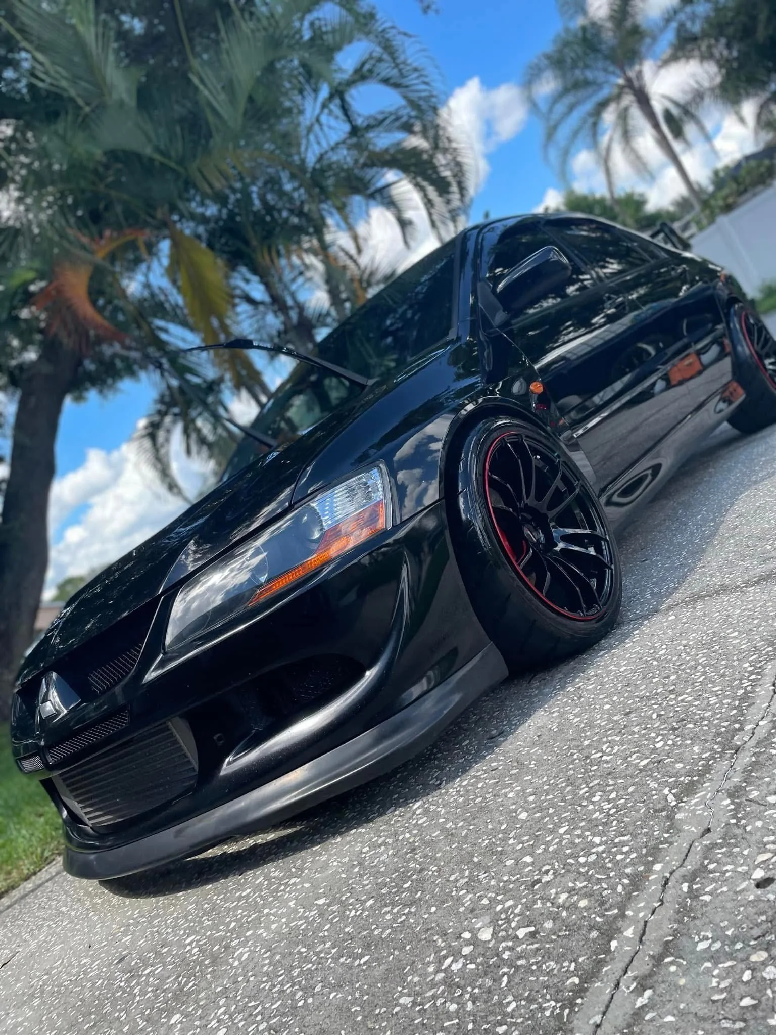 Black modified car with lowered stance and custom black rims with red accents, parked on a concrete driveway beneath palm trees with blue sky and white clouds in the background.