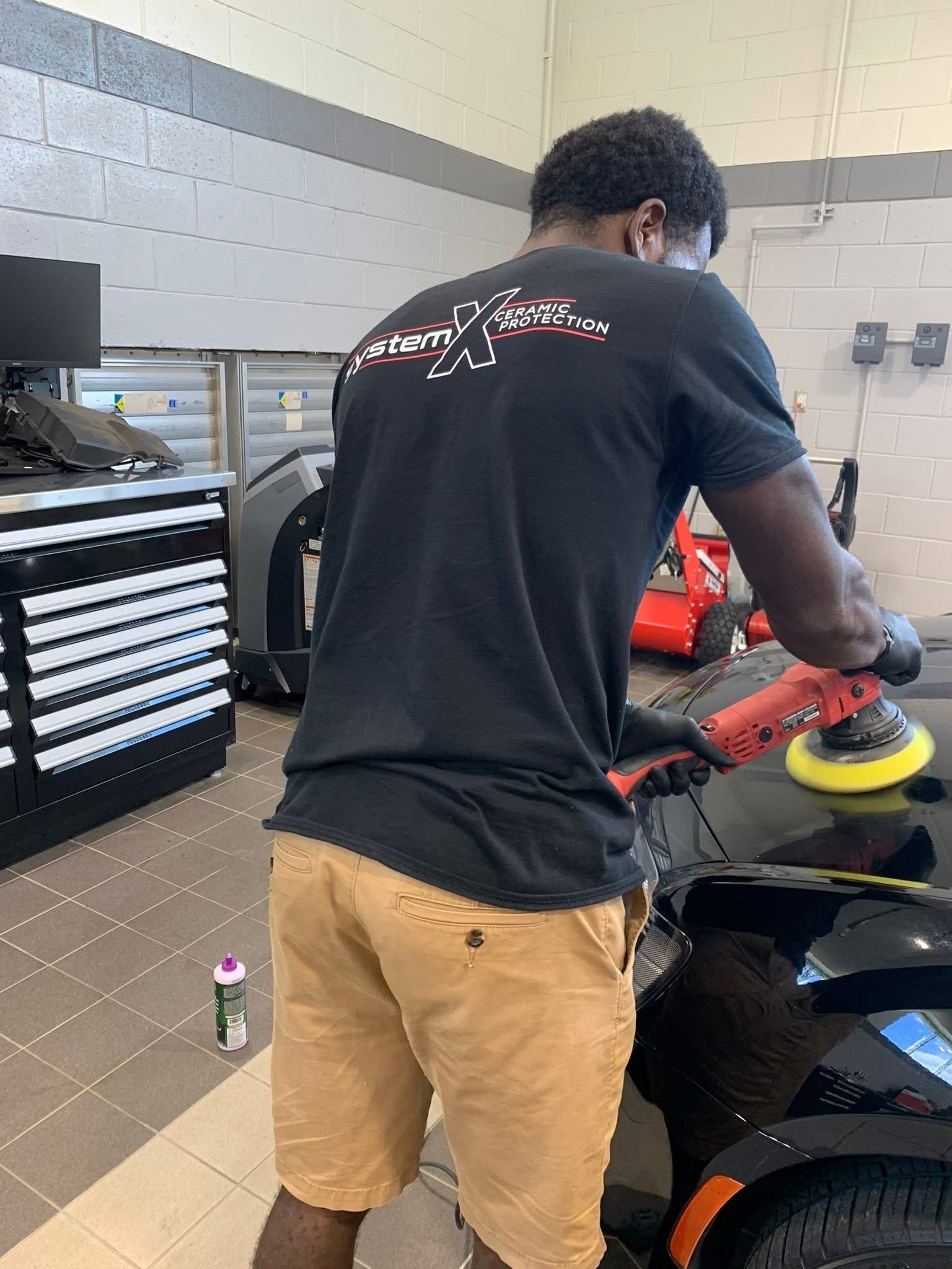 A person polishing a black car with a yellow and red power buffer in a garage or workshop.
