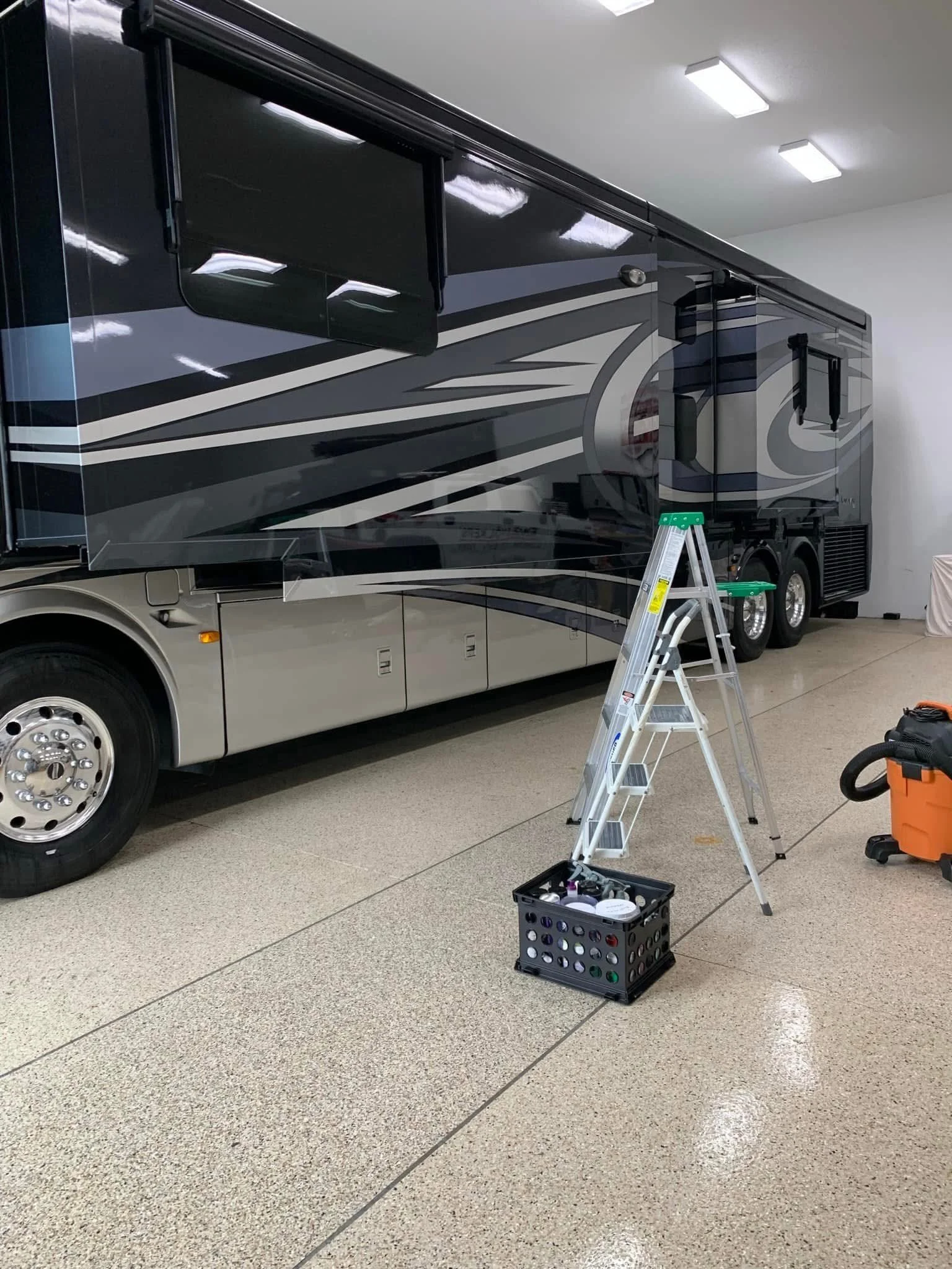 Interior view of a black and gray luxury RV with a ladder, toolbox, and cleaning supplies nearby, in a well-lit space.