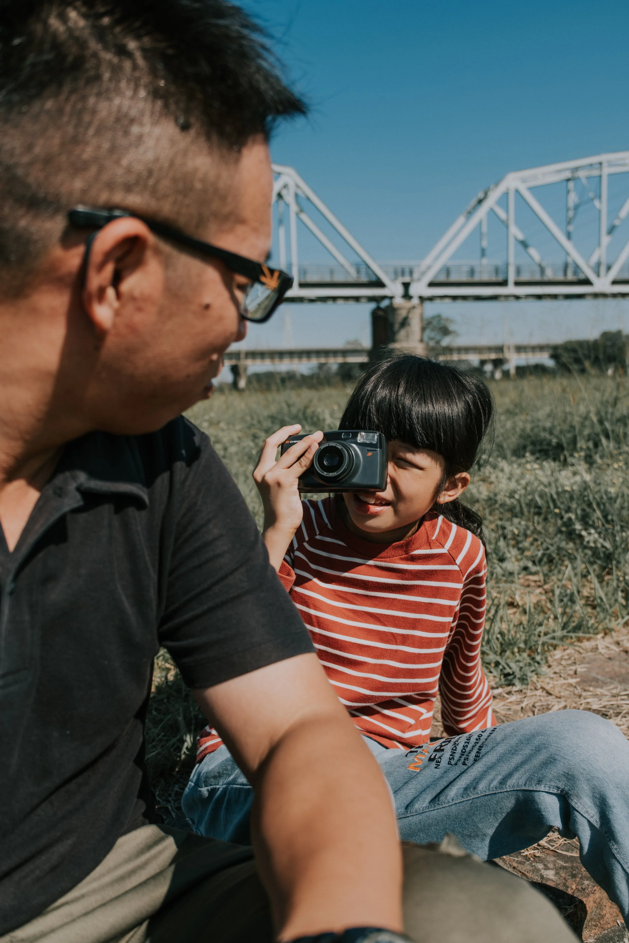 親子戶外寫真｜爸爸與孩子在橋邊草地相依偎，鐵橋為背景的溫情父子生活紀錄