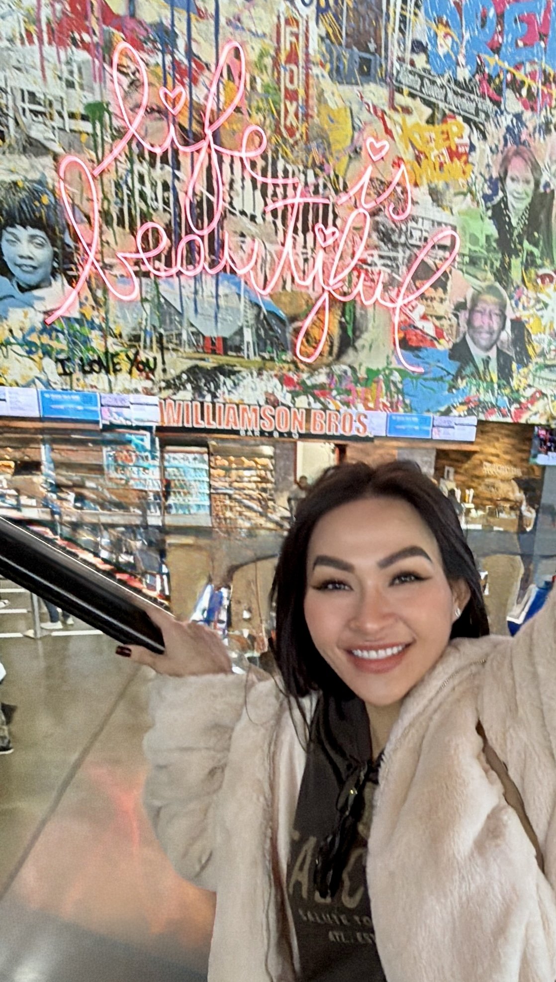 Trang T. Nguyen smiling under a neon ‘Life is beautiful’ sign on a colorful collage wall.